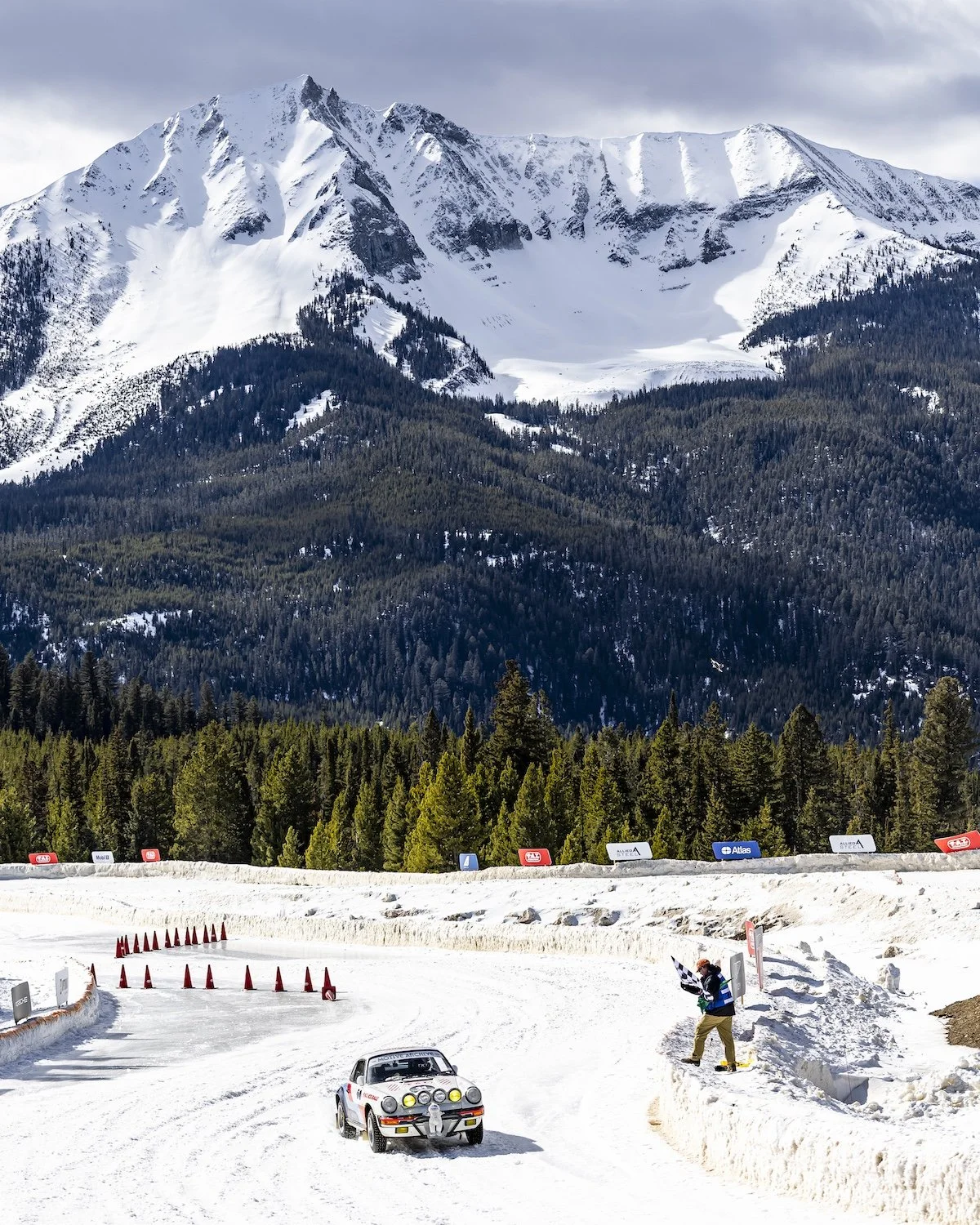 A rally car on a snow-covered mountain road with a person waving a checkered flag nearby, set against a backdrop of a forest and snow-capped mountains. F.A.T. ICE RACE 2026, Big Sky, Montana USA. Dan Lakatos Photo