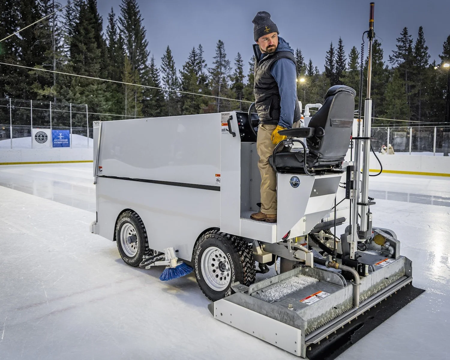 A person operating an ice-resurfacing machine on an outdoor ice rink surrounded by a forest of pine trees.