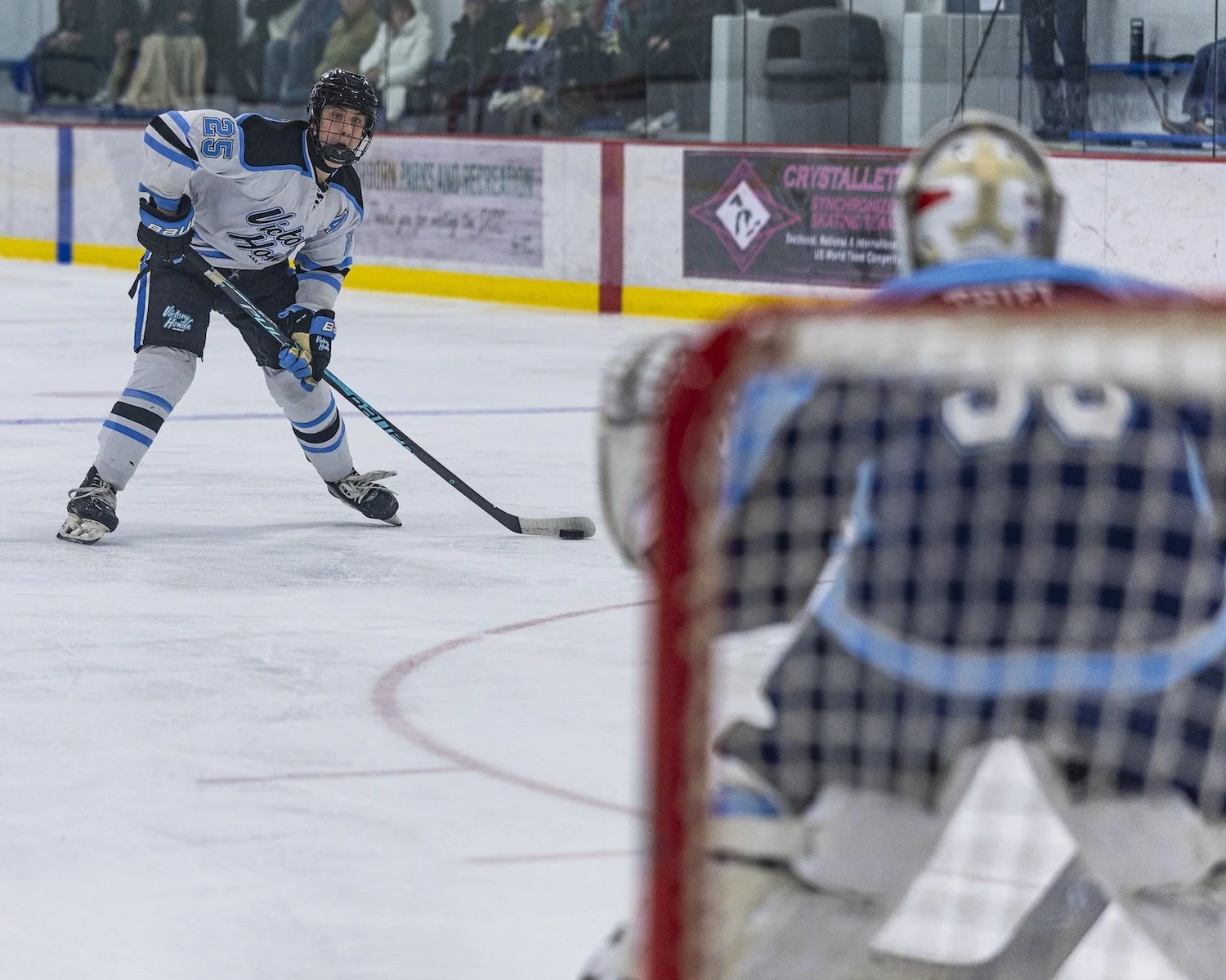 Hockey player in white and blue uniform prepares to take a shot at the goal while a goalie in checkered jersey and helmet watches from the net.
