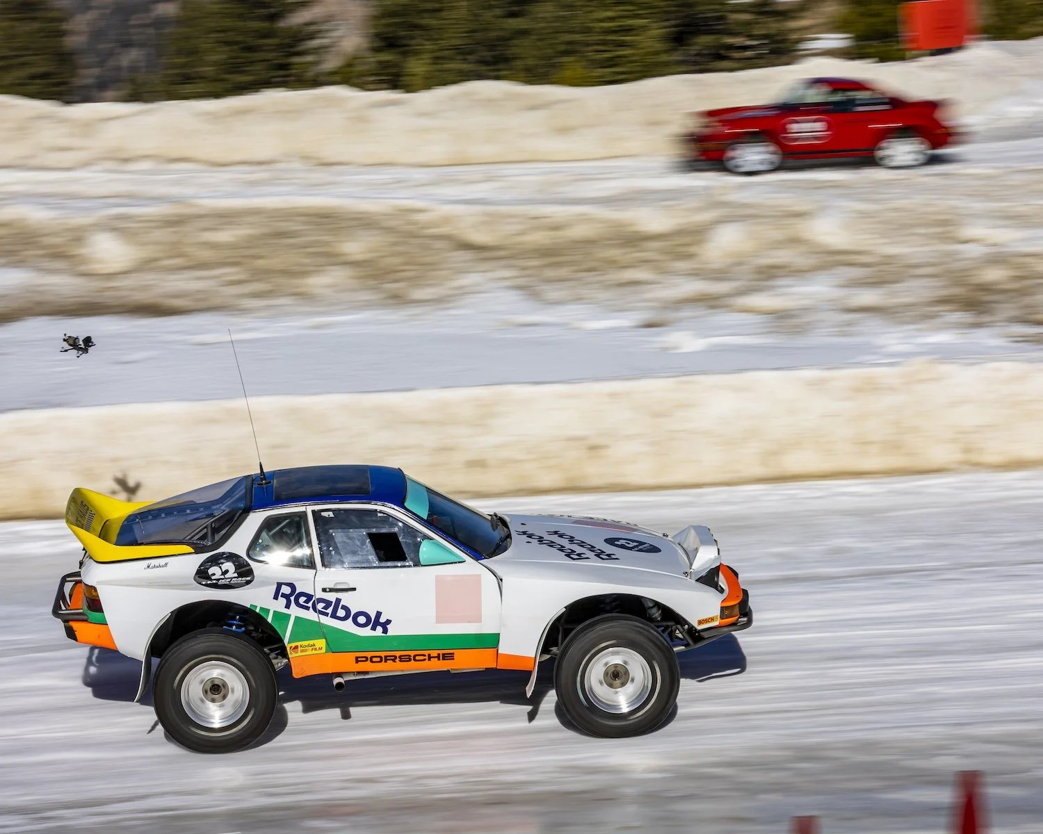 A classic Porsche race car with Reebok branding driving on a snow-covered track during a winter motorsport event, with a red car in the background. F.A.T. ICE RACE 2026, Big Sky, Montana USA. Dan Lakatos Photo