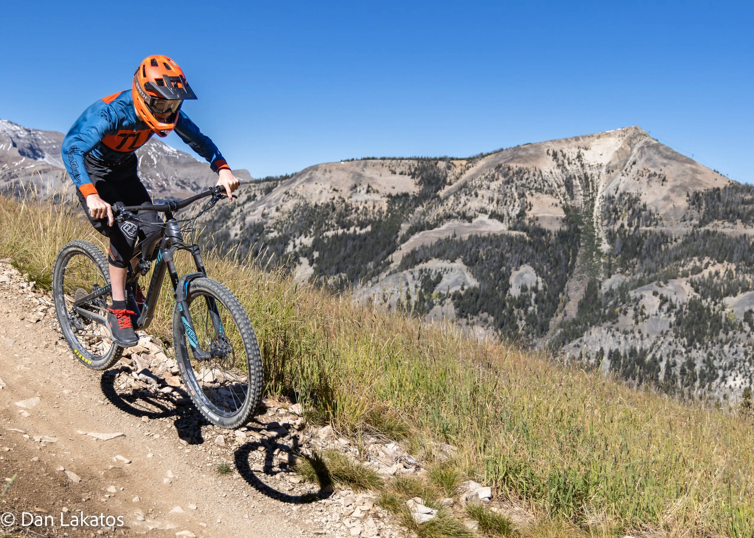 A person mountain biking downhill on a rocky trail in a mountainous terrain with clear blue sky and sparse trees in the background. Big Sky, MT, USA