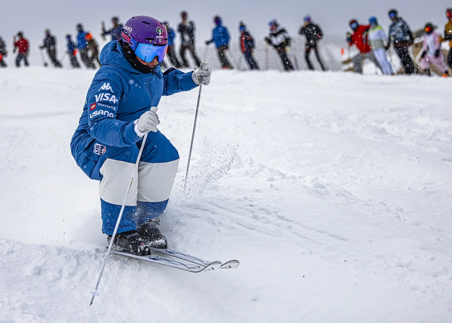 A snowboarder in a blue jacket and pants crouches on snow with skis and poles, wearing a purple helmet and goggles, with a crowd of people in winter clothing in the background.