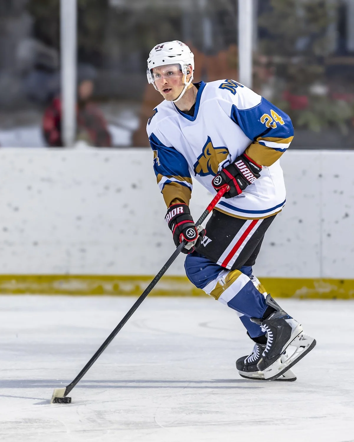 A hockey player in a white and blue jersey with gold accents, wearing a white helmet, gloves, and skates, skating on the ice with a hockey stick in hand, focused on the puck.