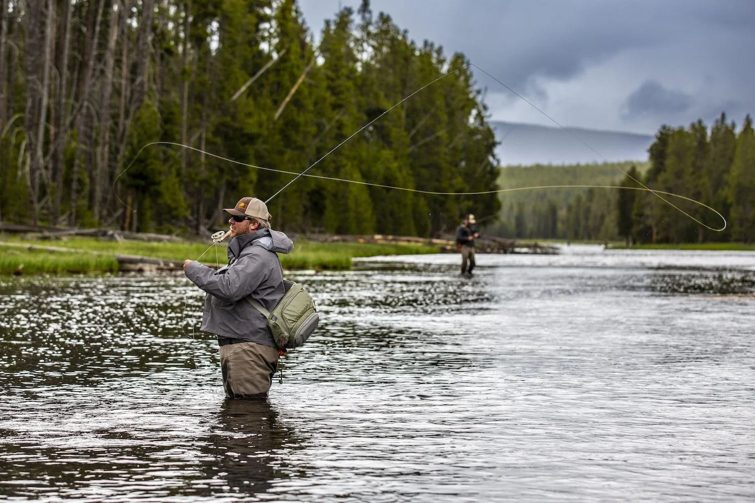 Two men fishing in a river surrounded by trees, with cloudy skies overhead. Yellowstone National Park, Wyoming, USA