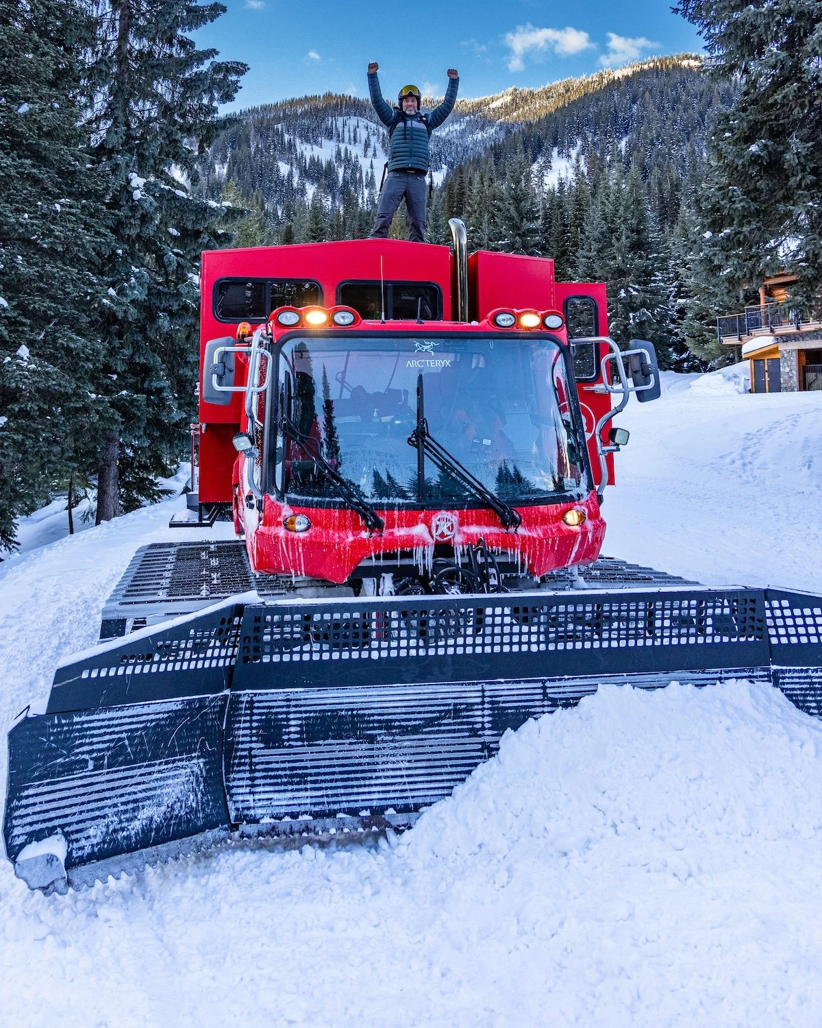 A person standing on top of a red snowcat machine in a snowy mountain landscape, raising their arms in celebration, with a forest and mountains in the background.