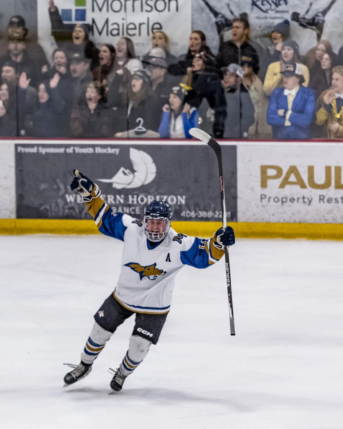 An ice hockey player celebrating on the ice rink with arms raised and a big smile, holding a hockey stick, wearing a white and blue jersey with a bird logo.
