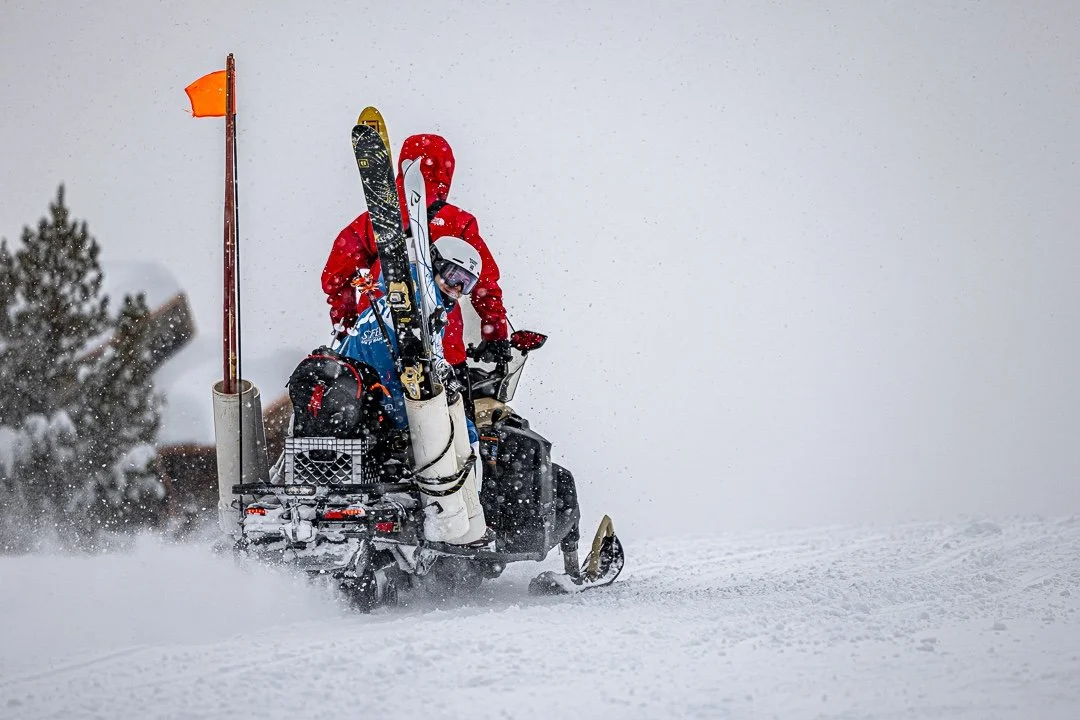 Person in red winter gear riding a snow scooter with skis, carrying gear, snow falling around them.