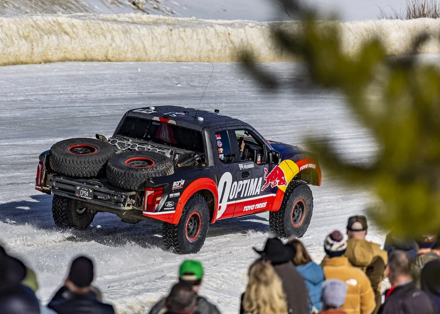 An off-road race truck with sponsorship decals driving on snow with spectators watching from the side. F.A.T. ICE RACE 2026, Big Sky, Montana USA. Dan Lakatos Photo
