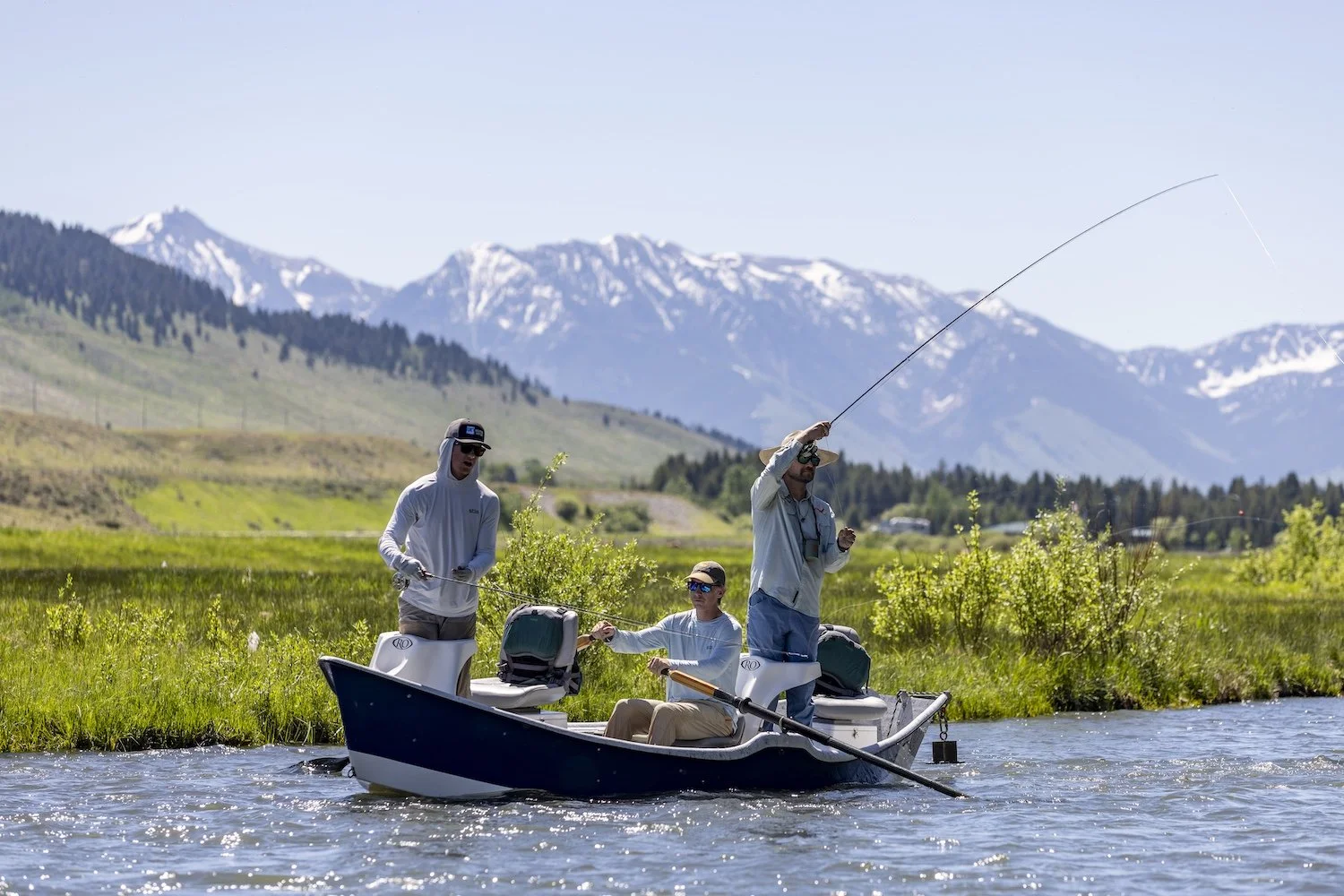 Three men fishing on a boat in a river with green banks, mountains in the background, and snowy peaks under a clear blue sky. Madison River, Montana, USA