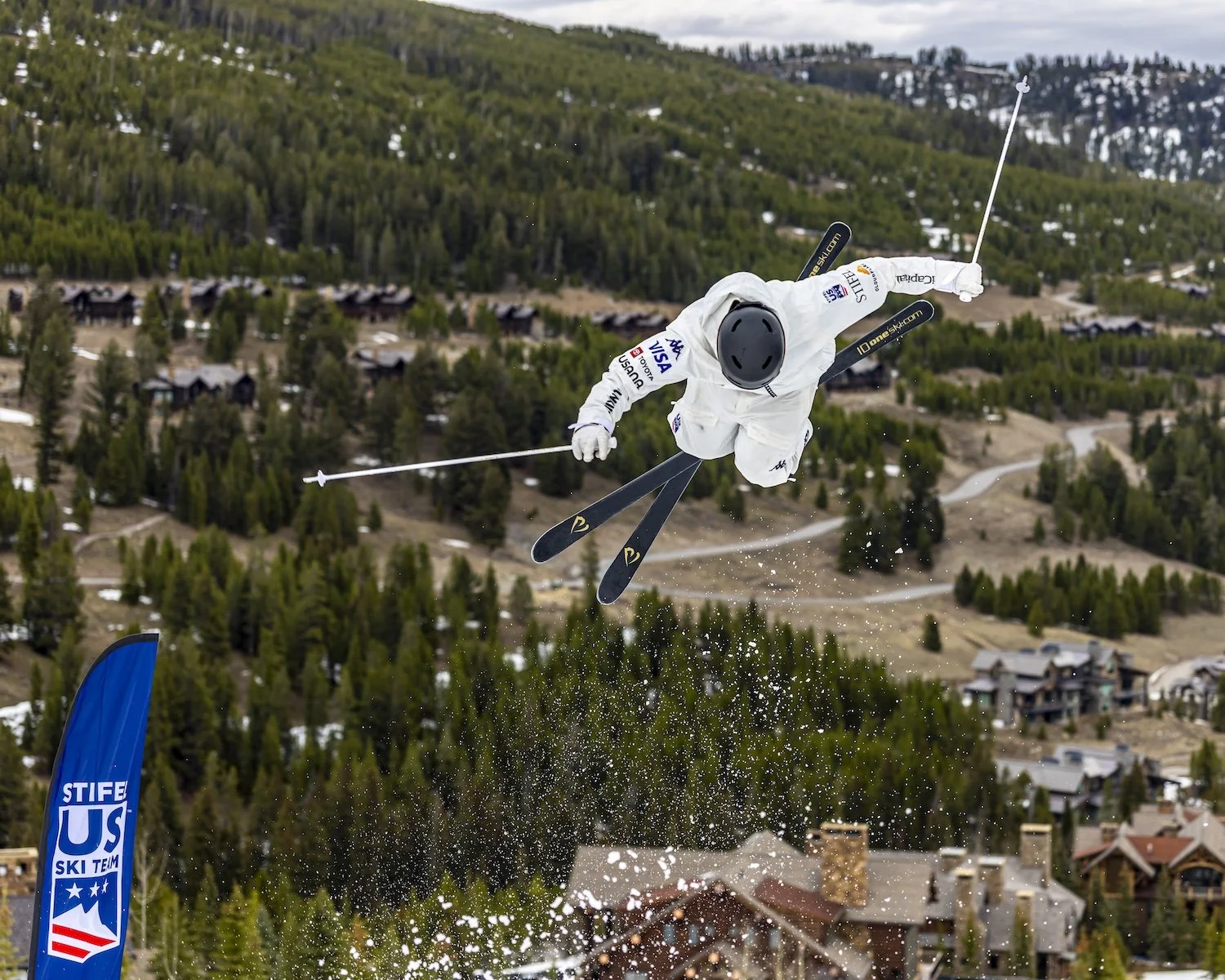 A skier in white gear and a black helmet performing an aerial trick against a mountainous landscape with trees and houses in the background.