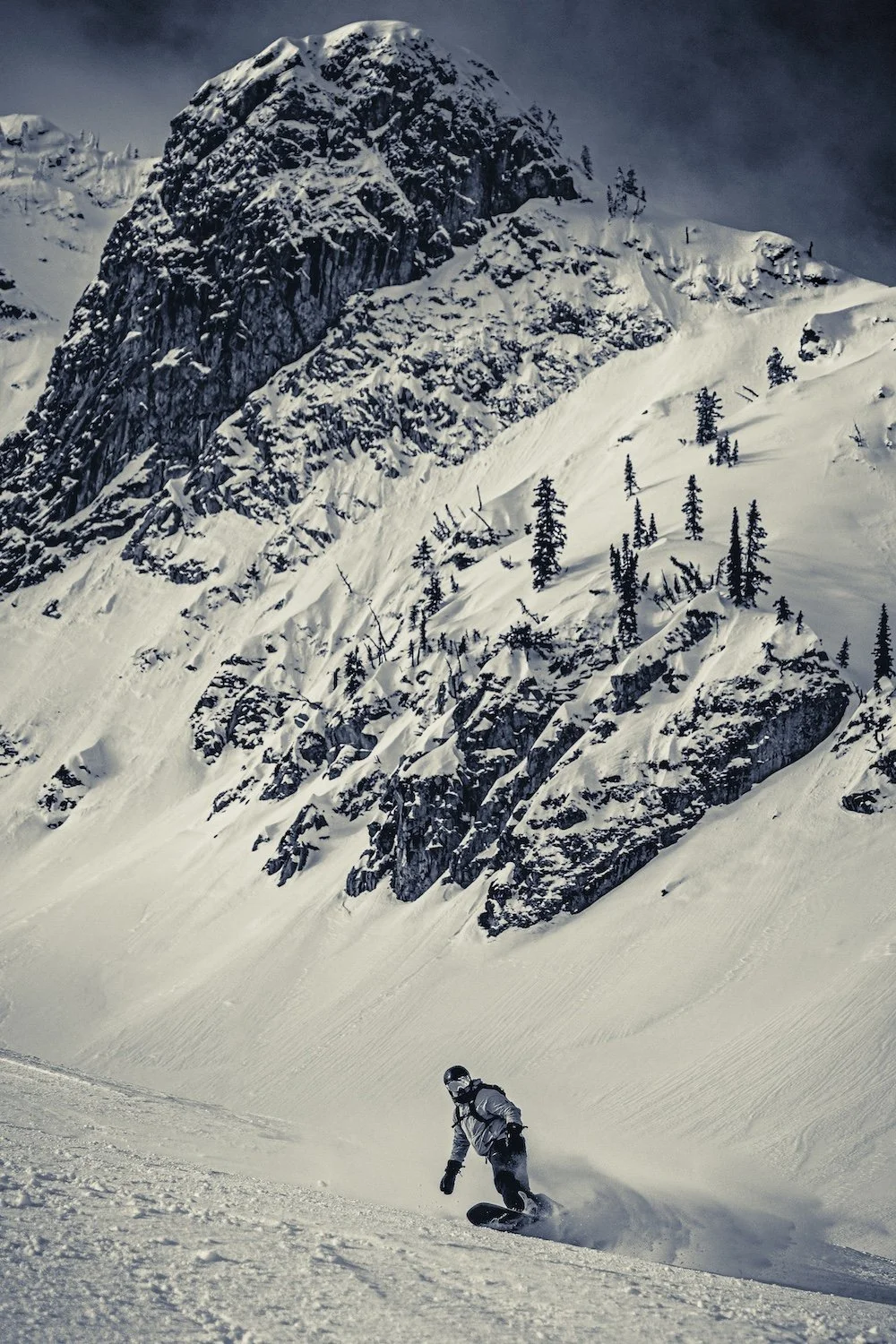 A person snowboarding down a snowy slope in a mountainous, forested area with large rock formations and evergreen trees.