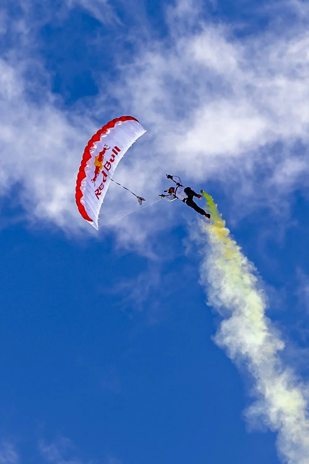 A skydiver with a Red Bull parachute descending through a blue sky with scattered clouds, trailing yellow-green smoke. F.A.T. ICE RACE 2026, Big Sky, Montana USA. Dan Lakatos Photo