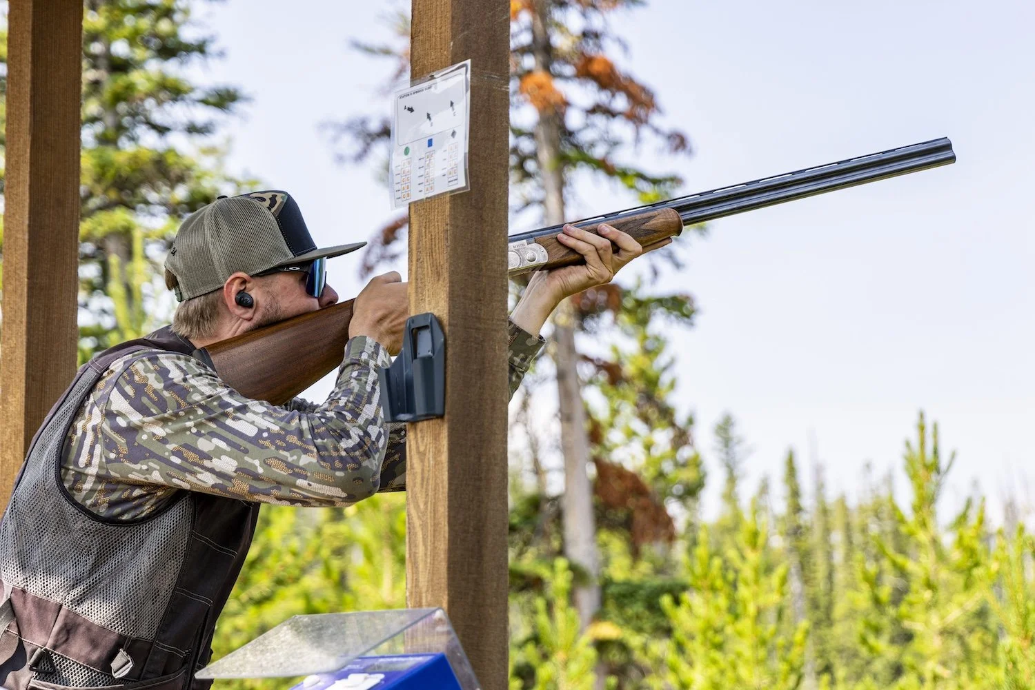 Man aiming a shotgun at a shooting range, using a wooden support for stability, surrounded by trees. Big Sky, Montana, USA