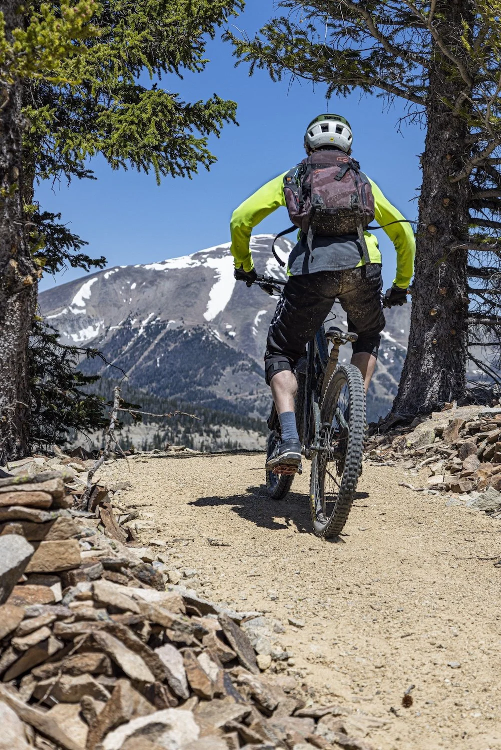 A person mountain biking on a dirt trail in a mountainous forest area, with snow-capped peaks in the background, wearing a helmet, bright yellow jacket, and carrying a backpack. Big Sky, MT, USA