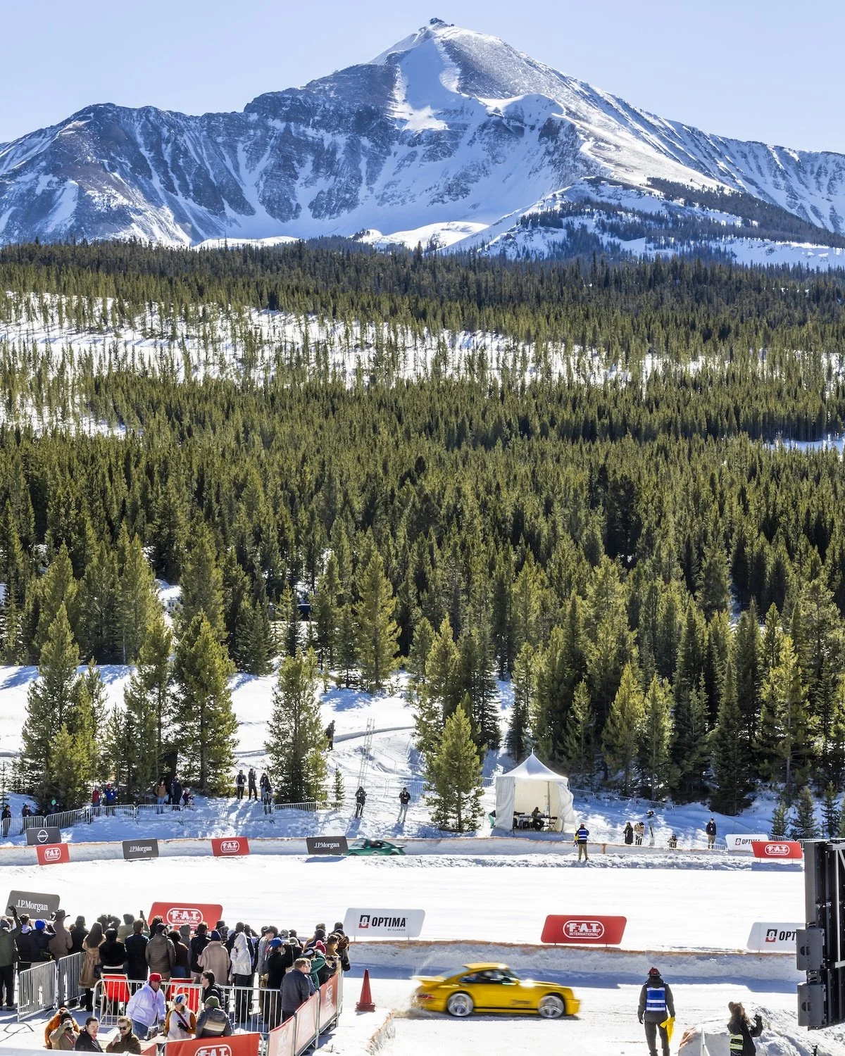 Yellow race car speeding on snow-covered track with spectators watching behind barriers, forested mountains and snow-capped peaks in the background. F.A.T. ICE RACE 2026, Big Sky, Montana USA. Dan Lakatos Photo