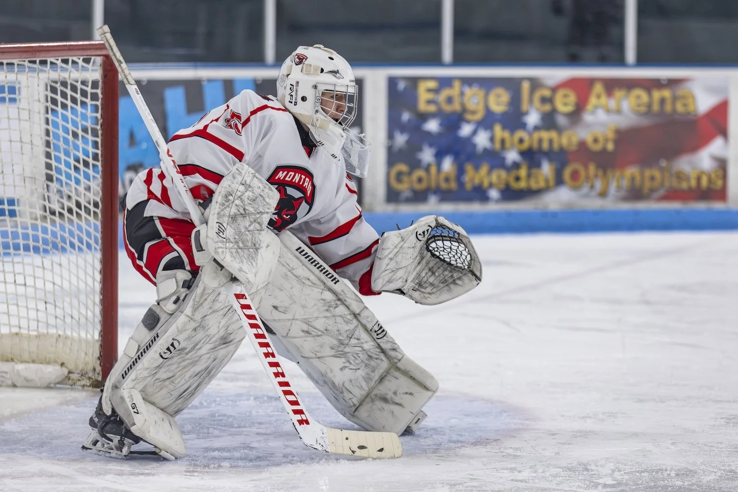 An ice hockey goaltender in a white and red uniform with a black and red emblem, squatting in front of the goal, preparing to make a save on the ice rink.