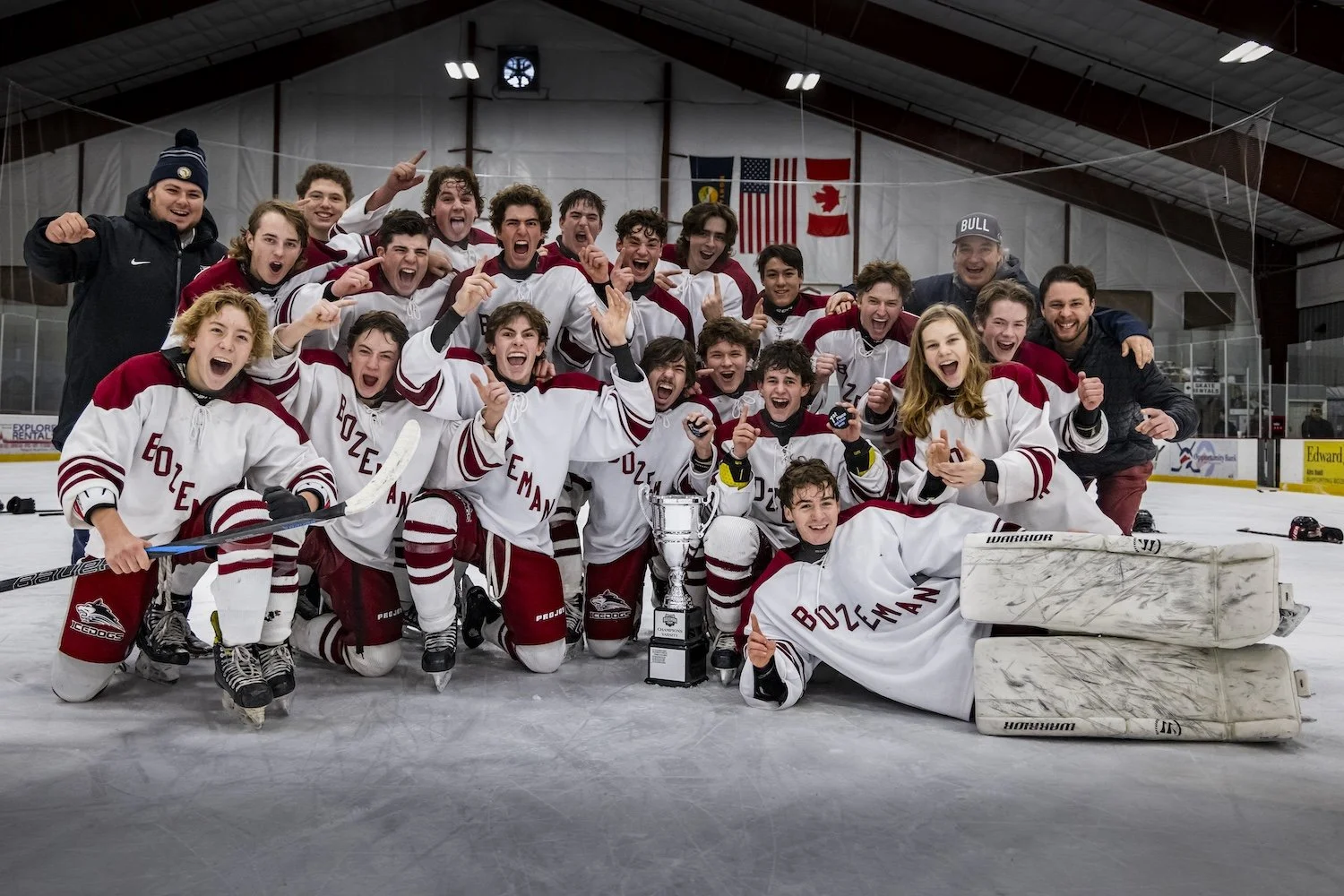 A group of young hockey players and coaches celebrating on the ice rink with a trophy, in front of an American flag and a Canadian flag, after a game or tournament.