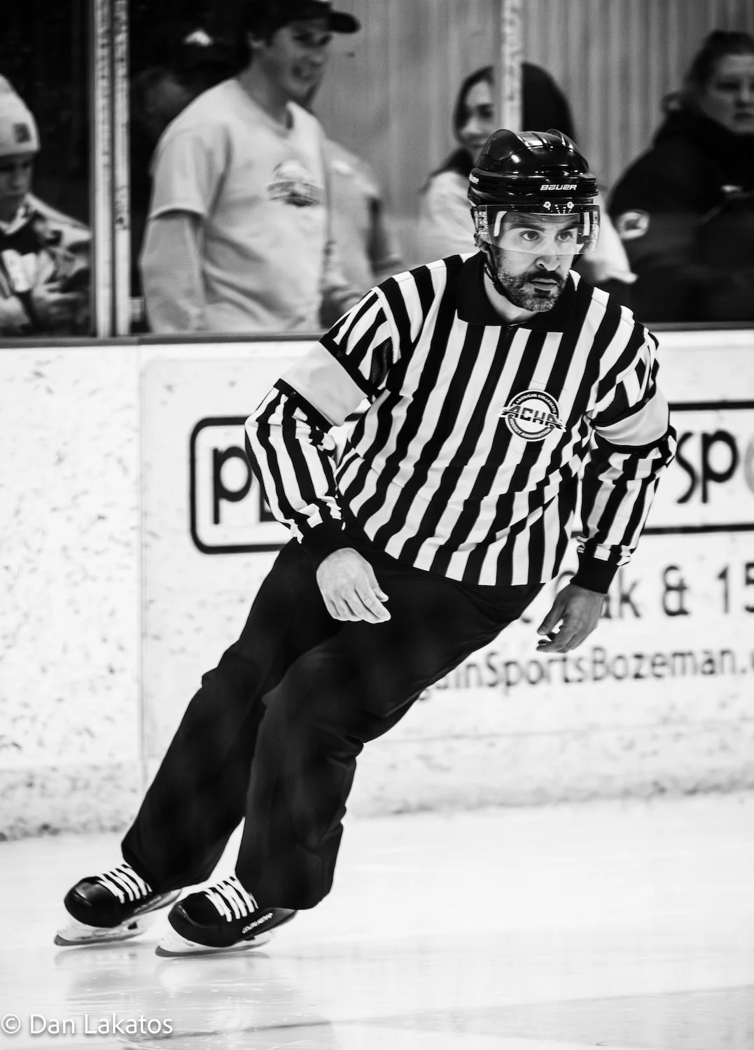 A hockey referee wearing a striped uniform and helmet, skating on an ice rink during a game, with spectators watching behind the glass.