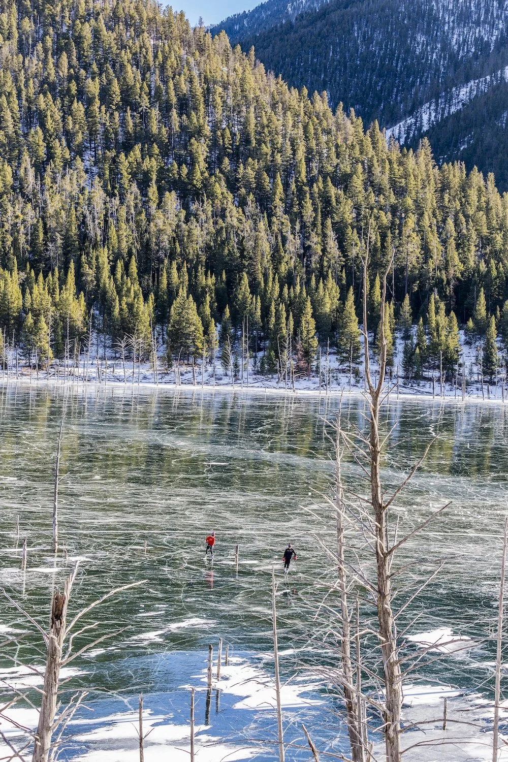 Two people walking on a frozen body of water surrounded by snow-covered trees and a densely wooded mountain in the background.