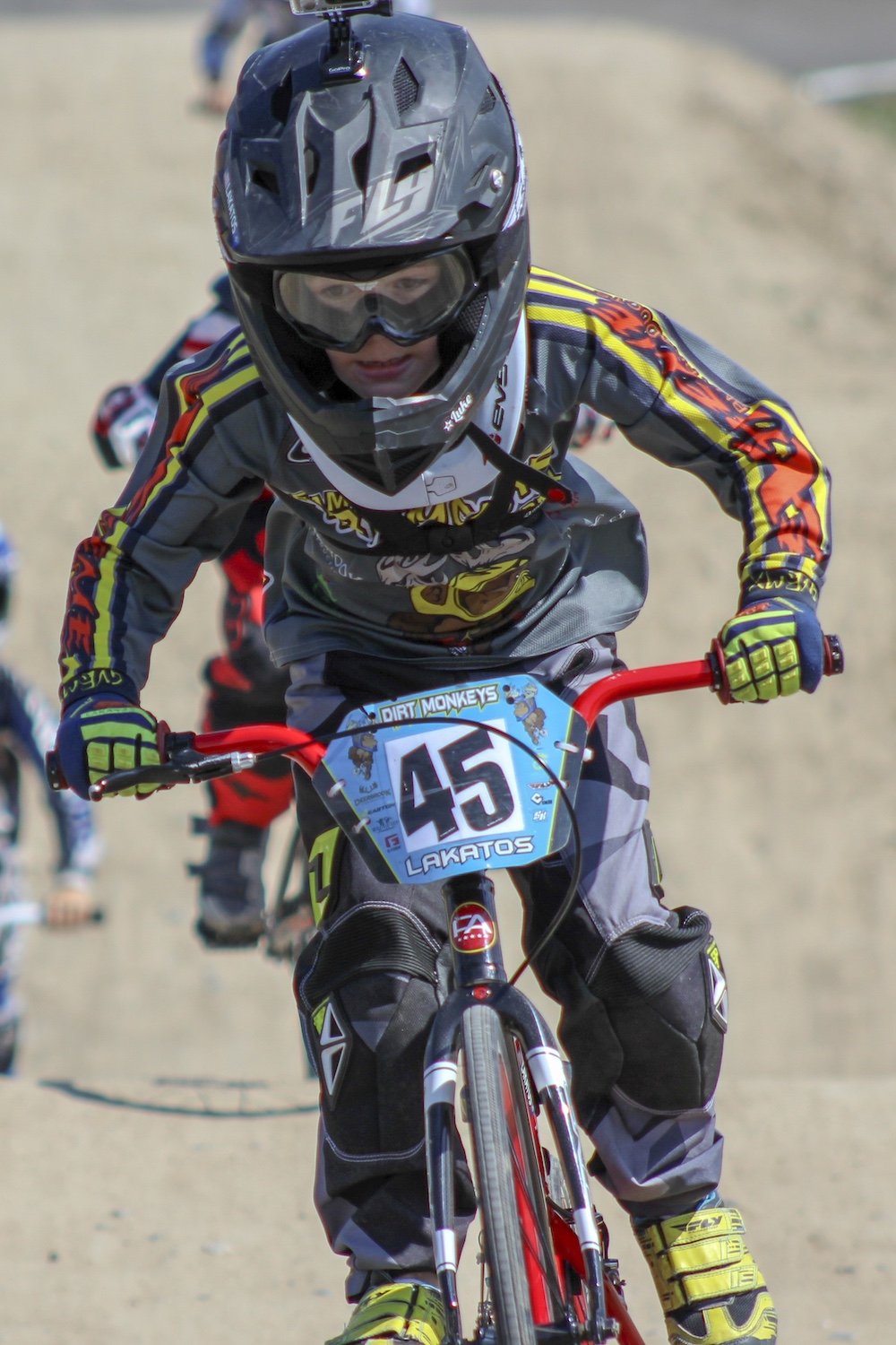 Young BMX rider in helmet and gear riding a dirt bike on a dirt track, with other riders in the background.