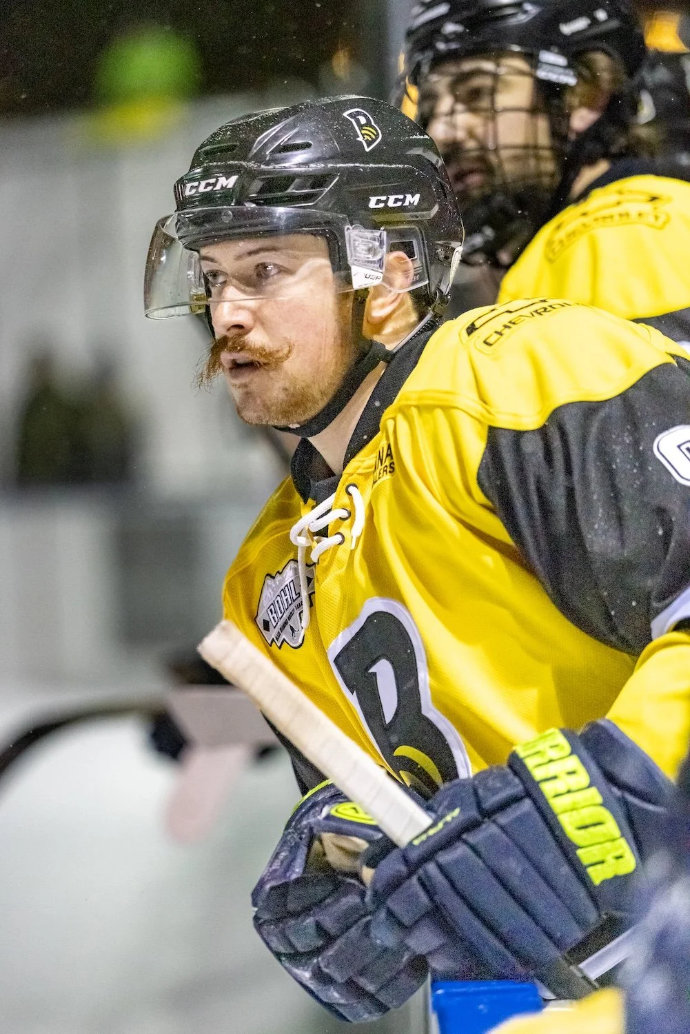 Ice hockey player in yellow jersey with black helmet and gloves, holding a hockey stick, on the ice during a game.