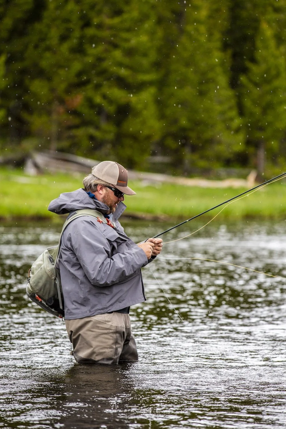 A man fishing in a river, wearing a gray jacket, beige pants, a hat, sunglasses, and a backpack, with trees in the background. Yellowstone National Park, Wyoming, USA