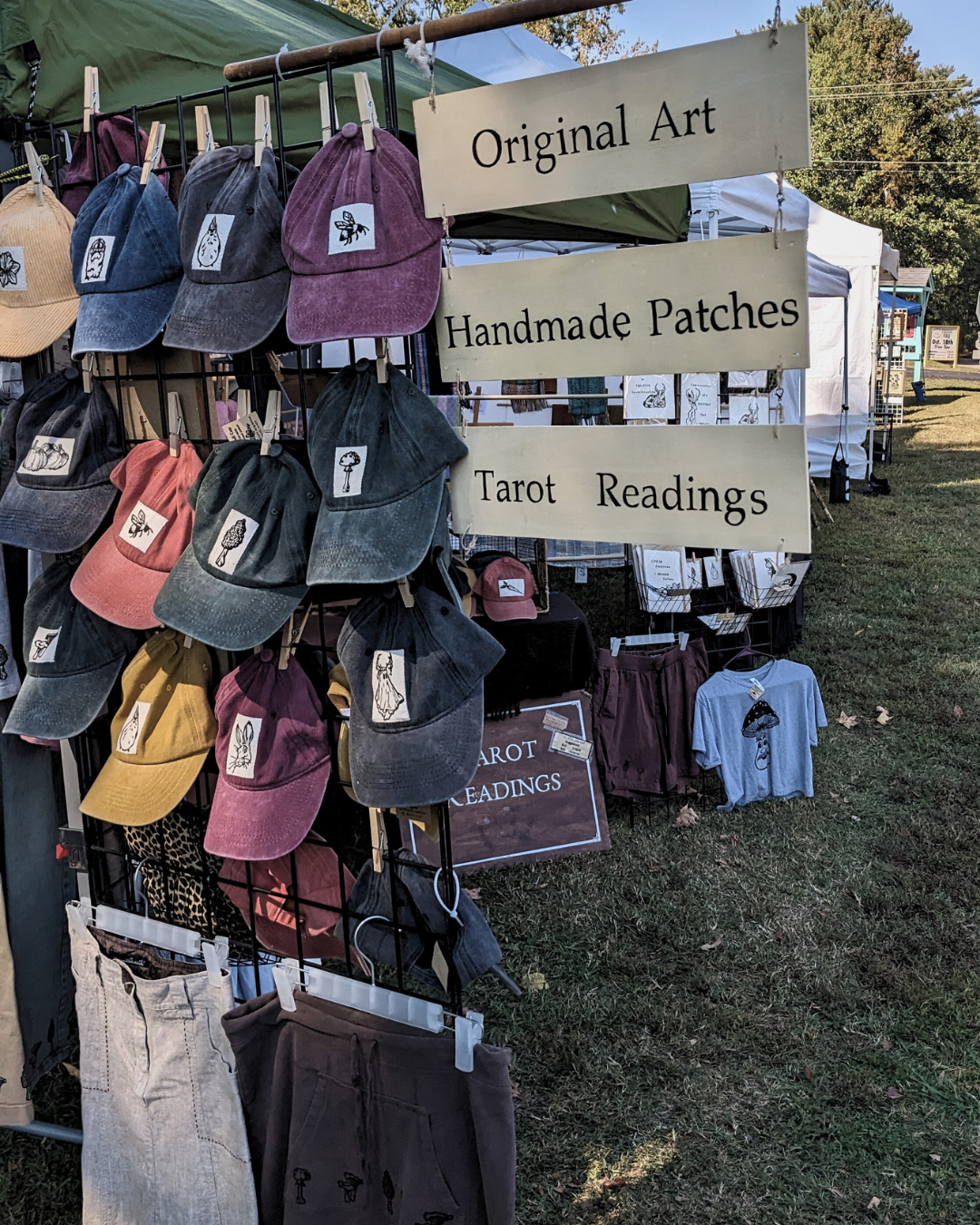 Art booth sign that reads "Original Art, Handmade Patches, Tarot Readings" with hats and other products in background.