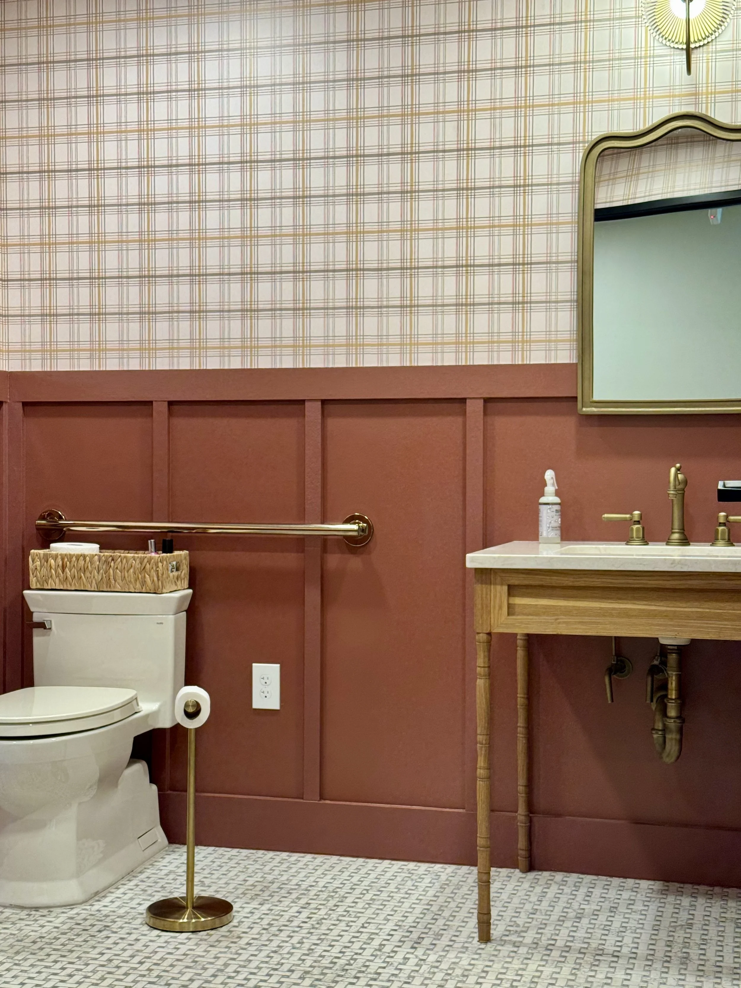 Bathroom with a toilet, a wooden sink vanity with a brass faucet, a mirror with a gold frame, a hand soap dispenser, and a hand-towel holder, with a patterned tile floor and plaid wallpaper.