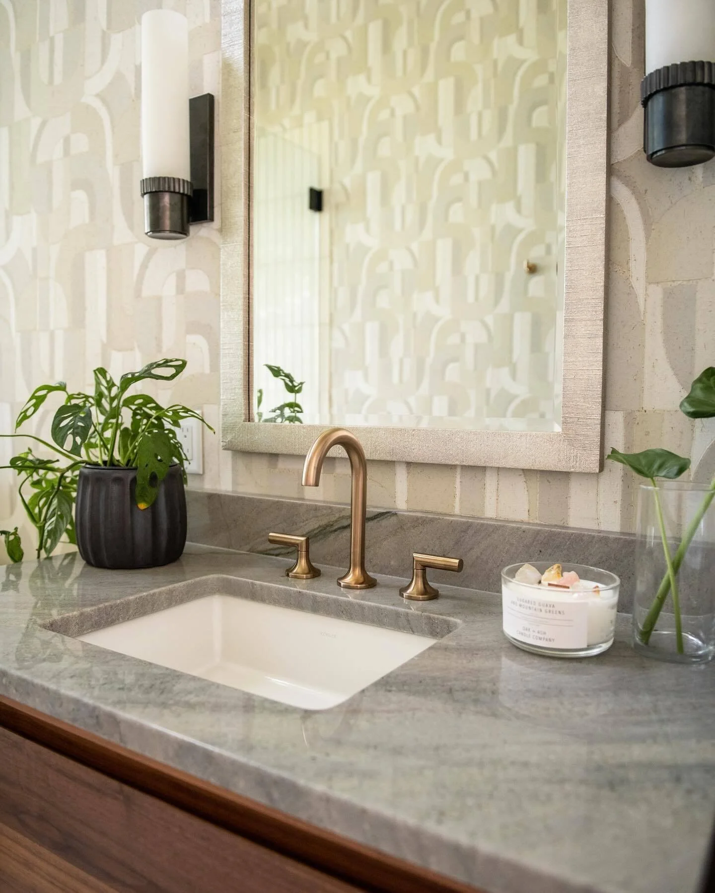 A bathroom vanity with a marble countertop, a white undermount sink, and a rose gold faucet. There are two potted plants, one with large leafs in a dark vase and another smaller plant in a clear vase. A lit candle with a white label and a small dish 