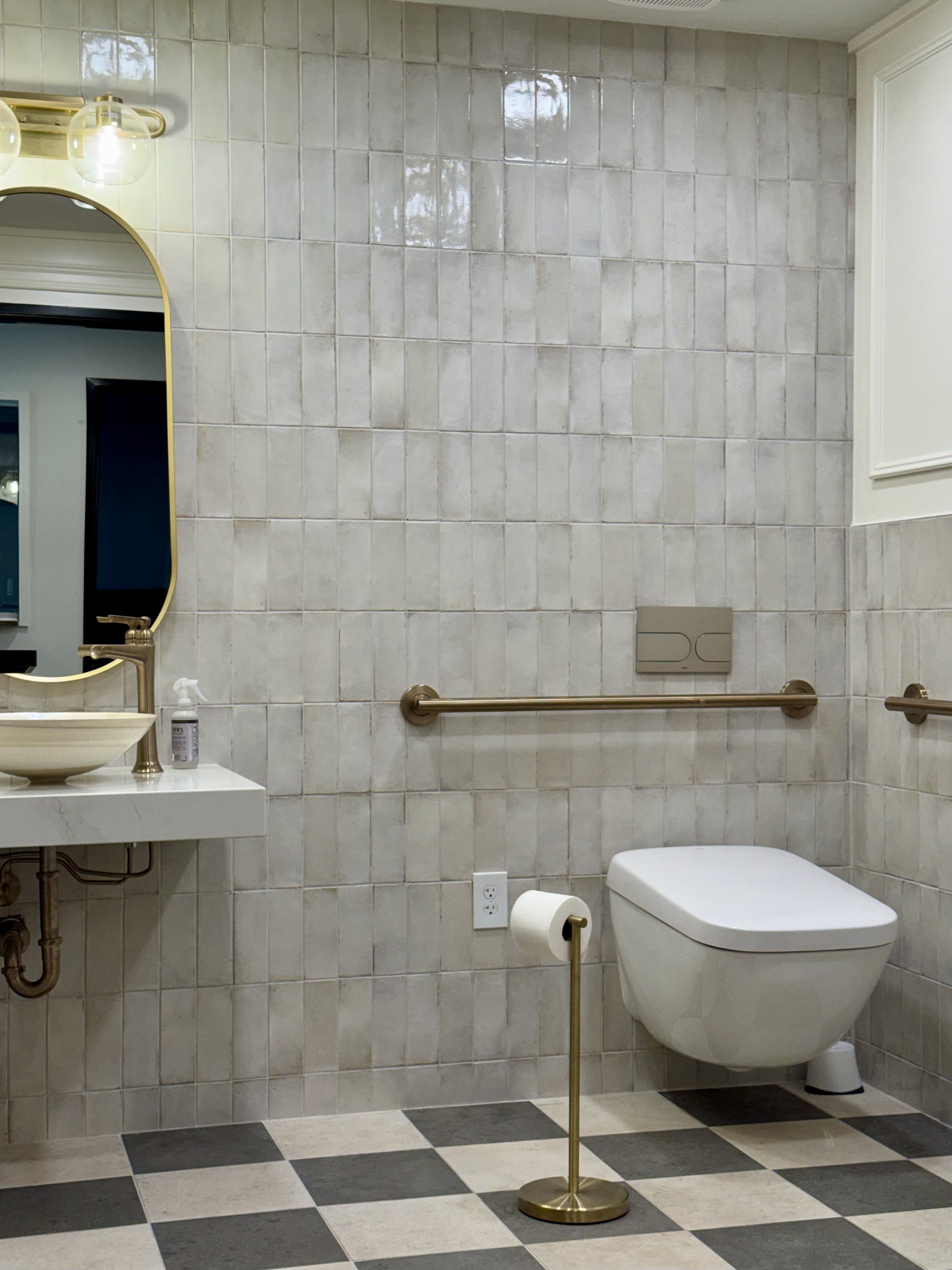 A bathroom with tiled walls and checkered black and white floor. It has a wall-mounted toilet, a small sink with gold fixtures, a mirror, and a towel bar. There's a bottle of hand sanitizer on the sink, and a gold-colored toilet paper holder with a roll of toilet paper.
