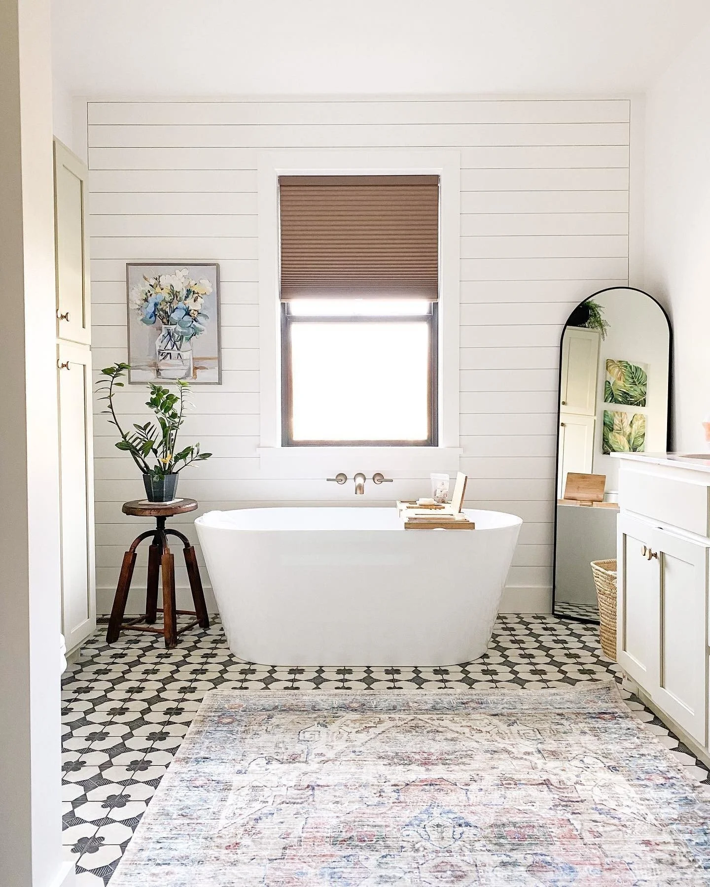 A modern bathroom with a freestanding bathtub, a window with a brown shade, black and white patterned floor tiles, a potted plant on a wooden stool, wall art of flowers, a tall mirror with plants, and white cabinetry.