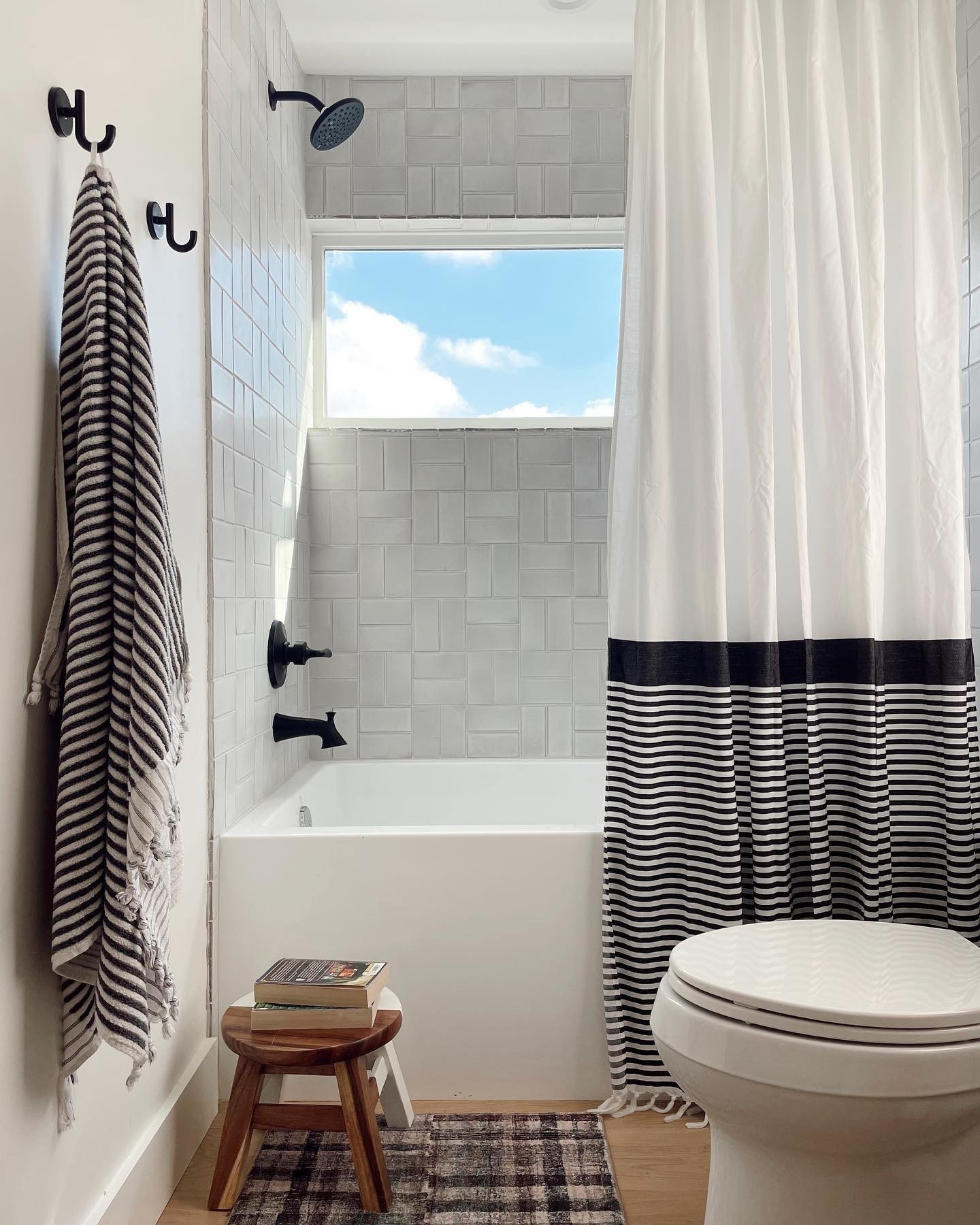 Bathroom with white tiled walls, black fixtures, a window with a view of blue sky and clouds, a shower with a dark showerhead, a black hook with a striped towel, a small wooden stool with books, a multicolored rug, a white curtain with black stripes,