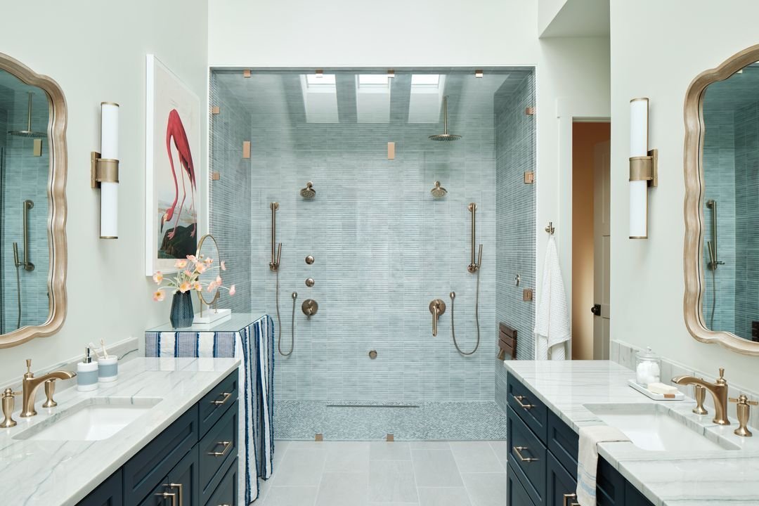 A modern bathroom with dual sinks, navy blue cabinets, marble countertops, and gold fixtures. Features a large glass-enclosed shower with dual showerheads.