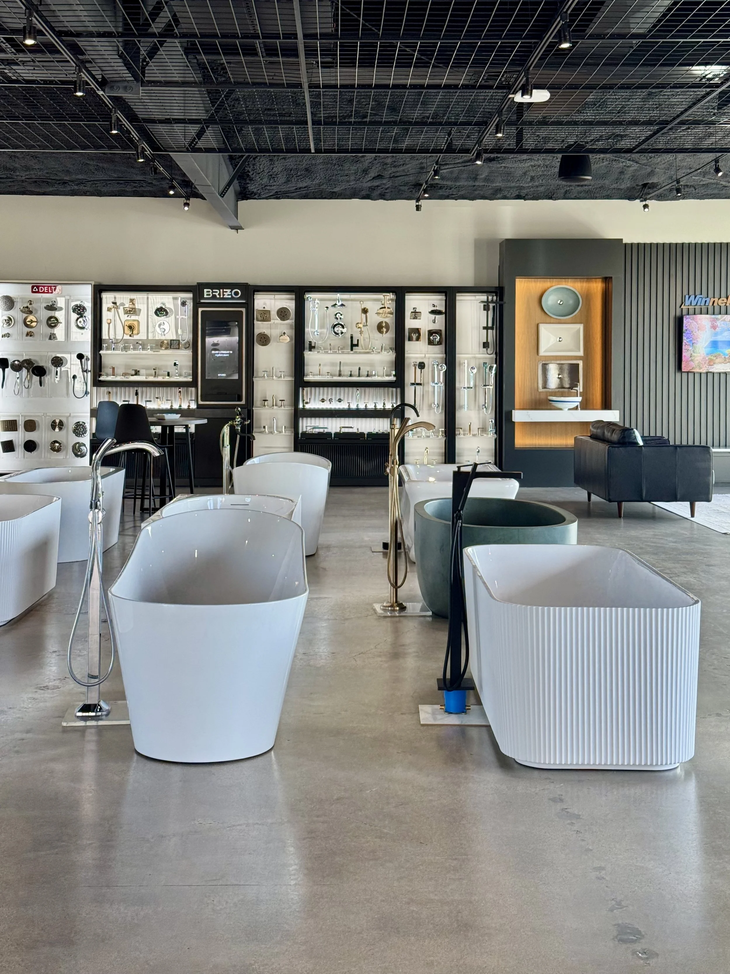 Showroom with modern bathroom fixtures and furniture, including white freestanding tubs, faucets, and sinks on display, with a black sofa and decorative wall panels.