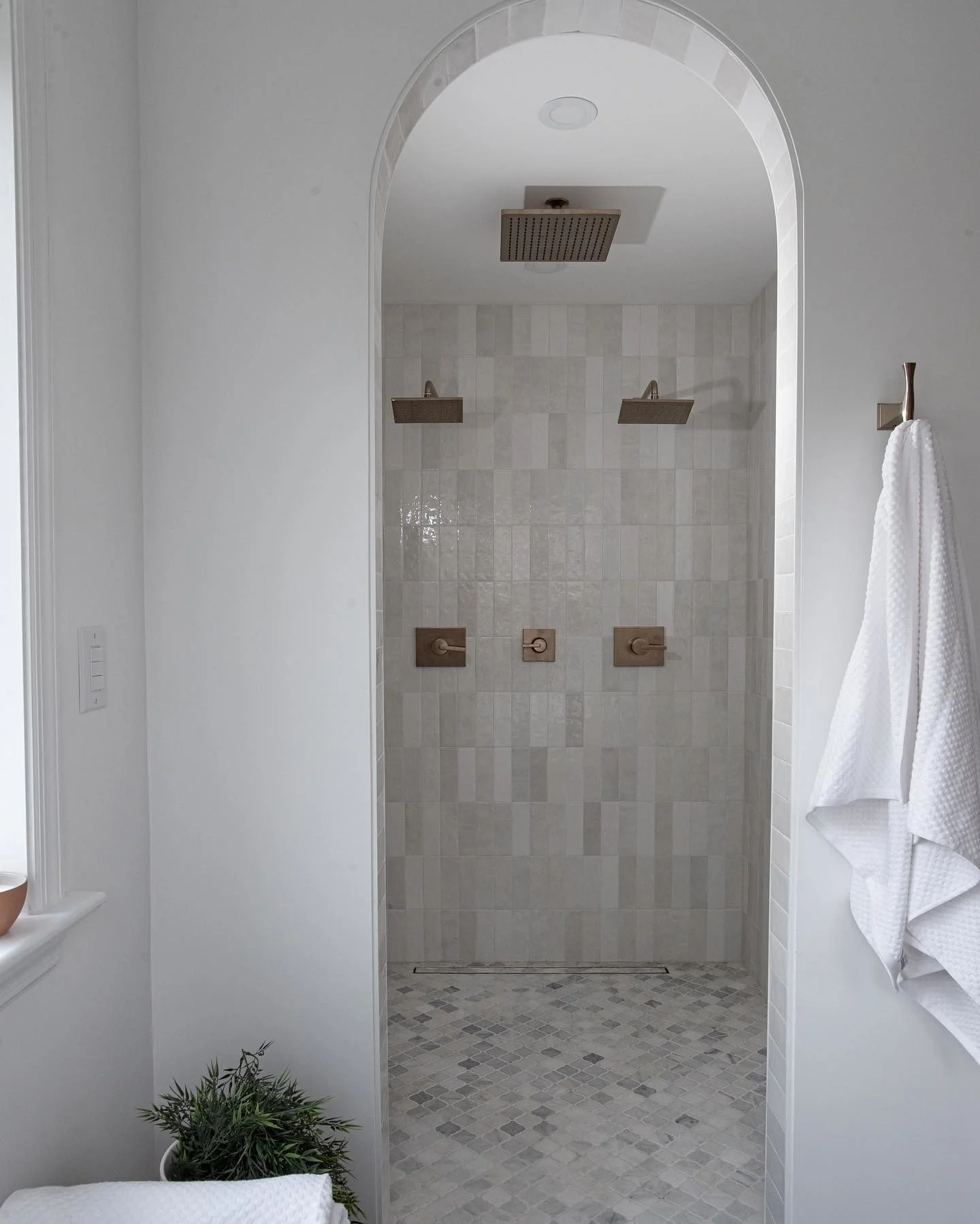 Clean modern shower with beige tiles, rain showerhead, two wall-mounted showerheads, and a twist valve, with white walls and a white towel hanging on a hook to the side.