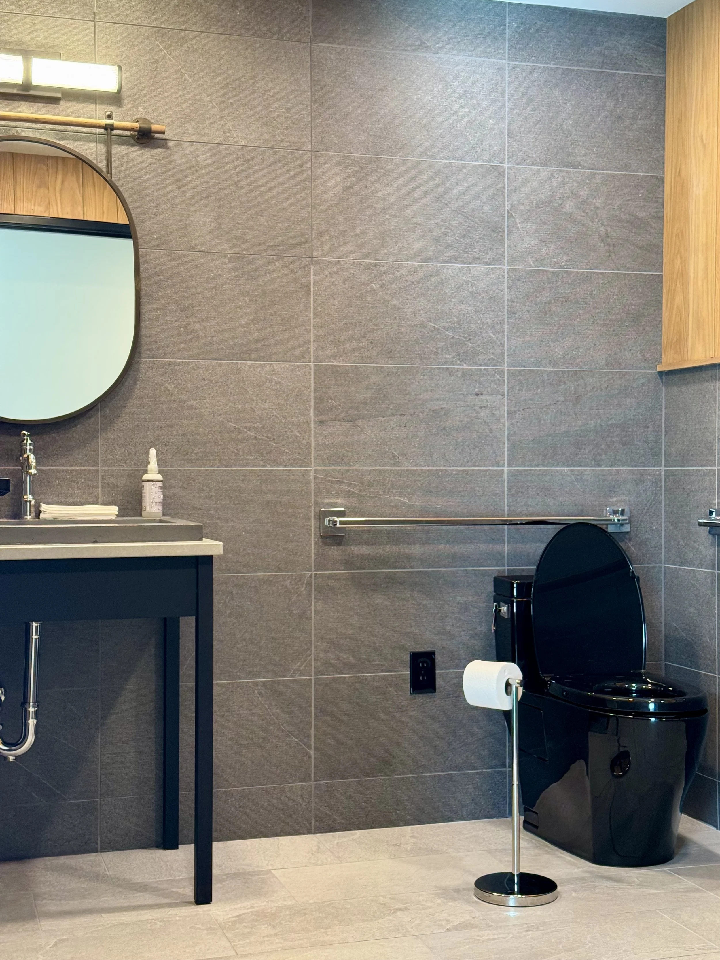 A bathroom with gray tiled wall, a black toilet with a closed lid, a toilet paper stand with a roll, a wooden vanity with a gray countertop, a mirror, a bottle of liquid soap, and a towel bar.