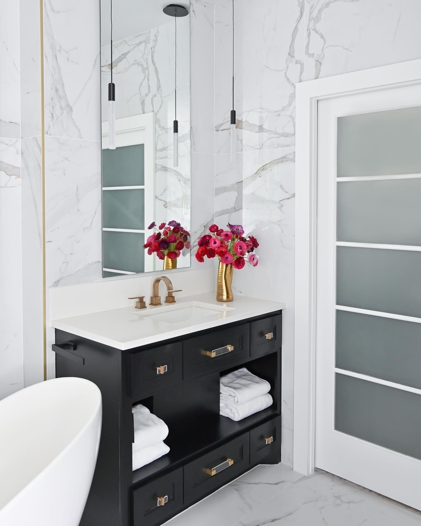 Modern bathroom with black vanity, white countertop, gold handles, and white towels. Wall and floor have marble design. Tall mirror with pendant lights, gold vase with pink and purple flowers, and frosted glass door are visible.