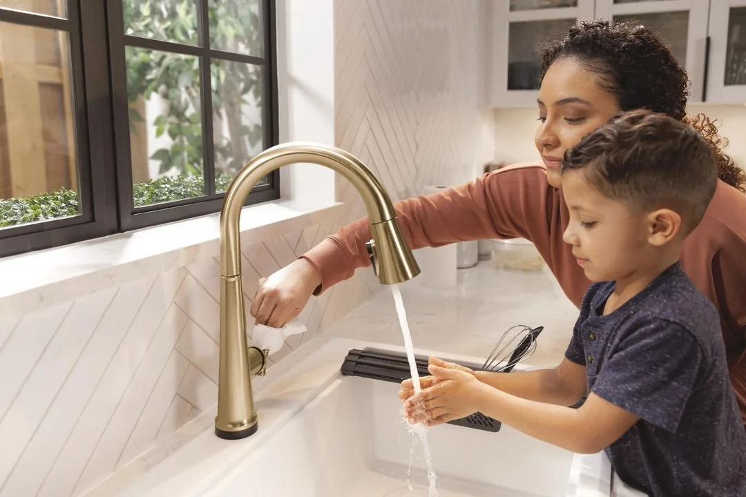 A woman helping a young boy wash his hands at a kitchen sink with a golden faucet, near a window with greenery outside.
