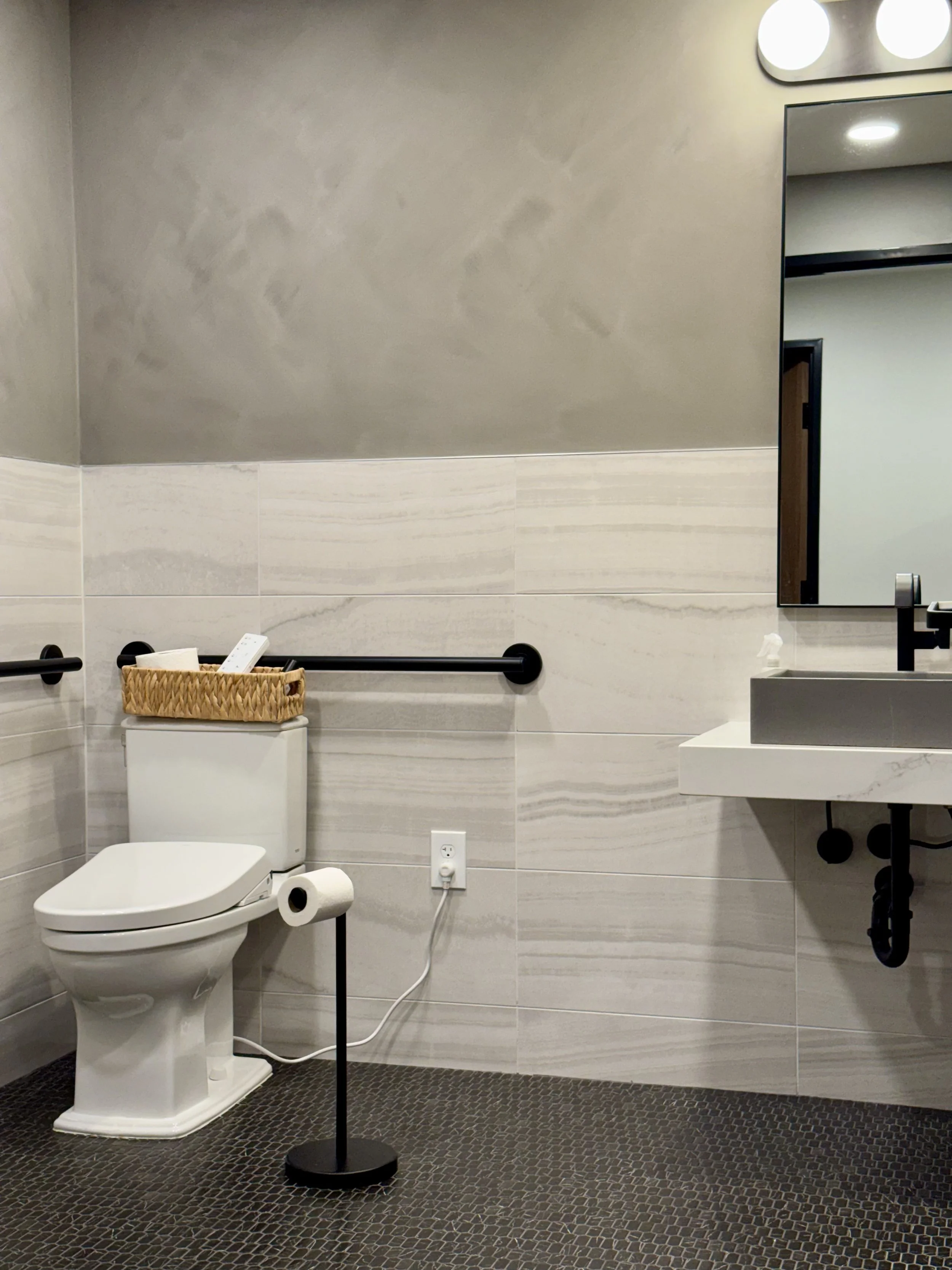 Bathroom with a white toilet, black railings, a mirror, black fixtures, a basket with toiletries, and a textured tile floor.