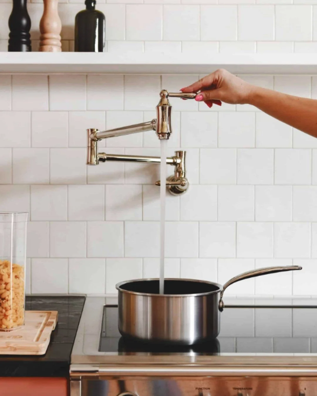 A hand turning on a kitchen faucet with water flowing into a stainless steel pot on a stove in a modern kitchen.