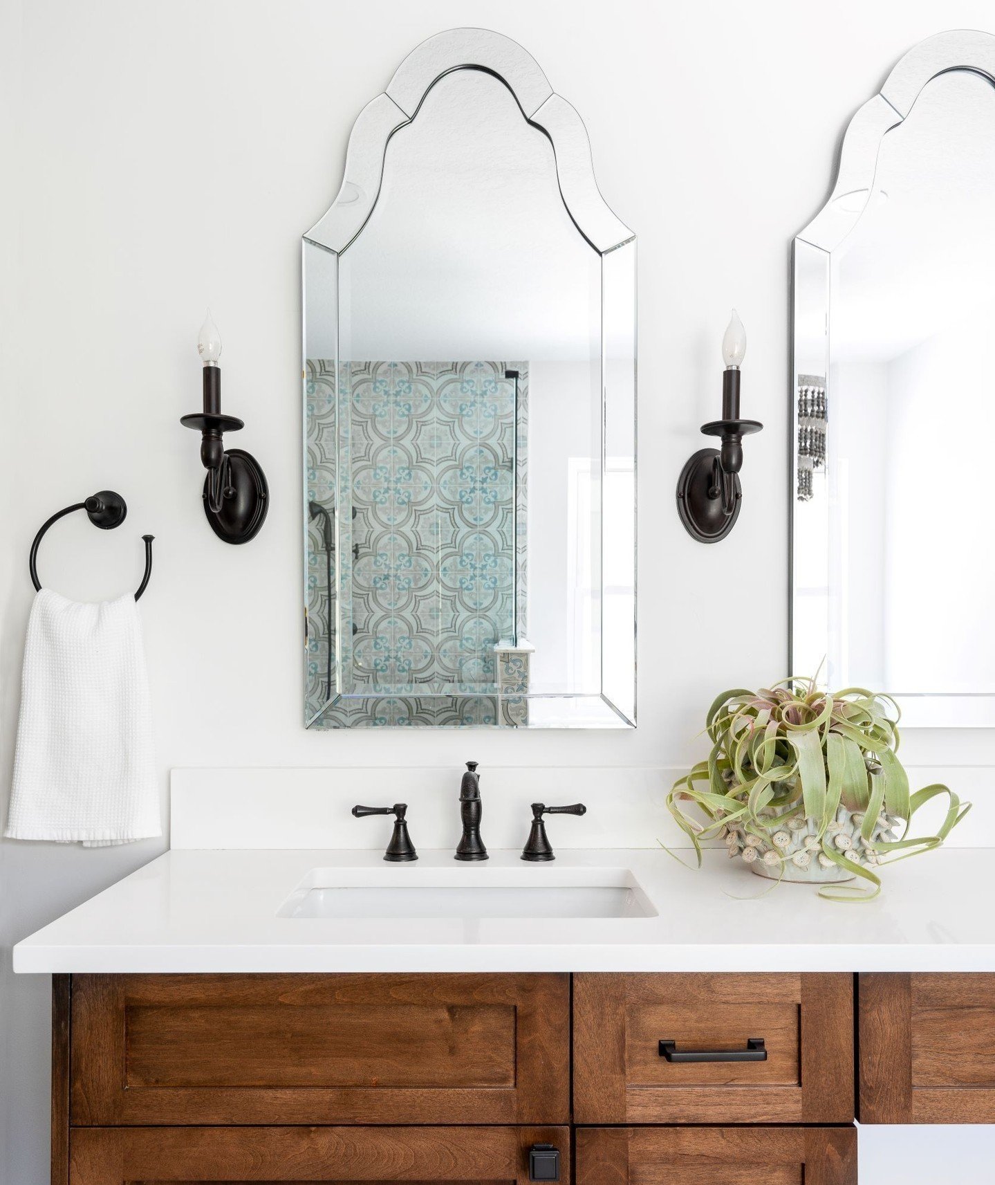 Bathroom vanity with white countertop, black faucet, wooden cabinet, and a mirror. Two black wall sconces with candles, a towel ring with white towel, and a potted plant are on the wall.