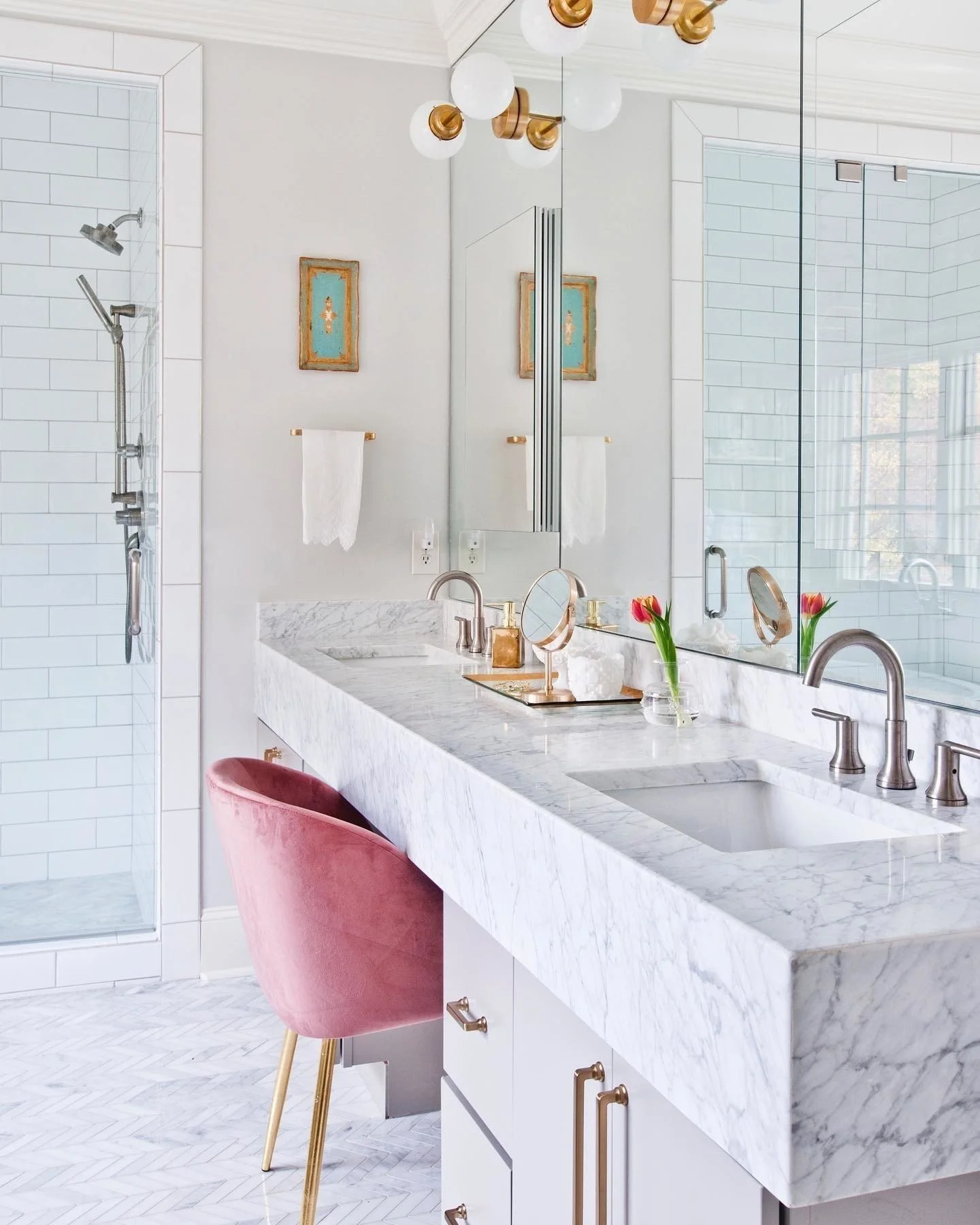 A modern bathroom featuring a white marble countertop with two sinks, gold and silver fixtures, and a large mirror. Pink velvet chair with wooden legs is placed in front of the countertop. Two small vases with pink tulips are on the counter. The bath