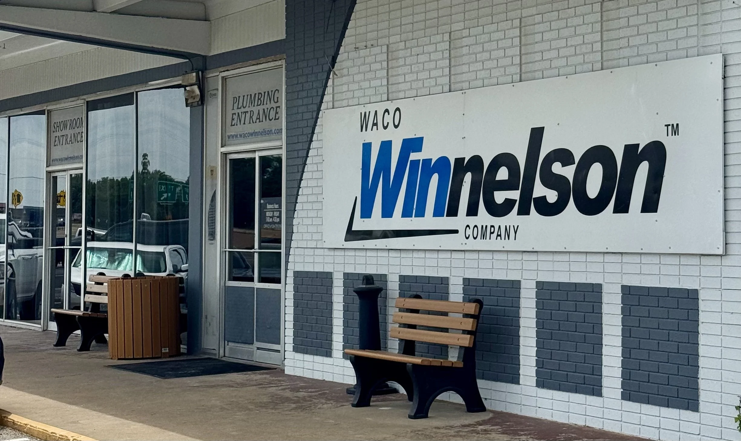 Exterior of a building with a large sign that reads "Waco Winelson Company." The building features glass doors and windows, with signs indicating entrances for the showroom and plumbing. There are benches and trash bins outside the building.
