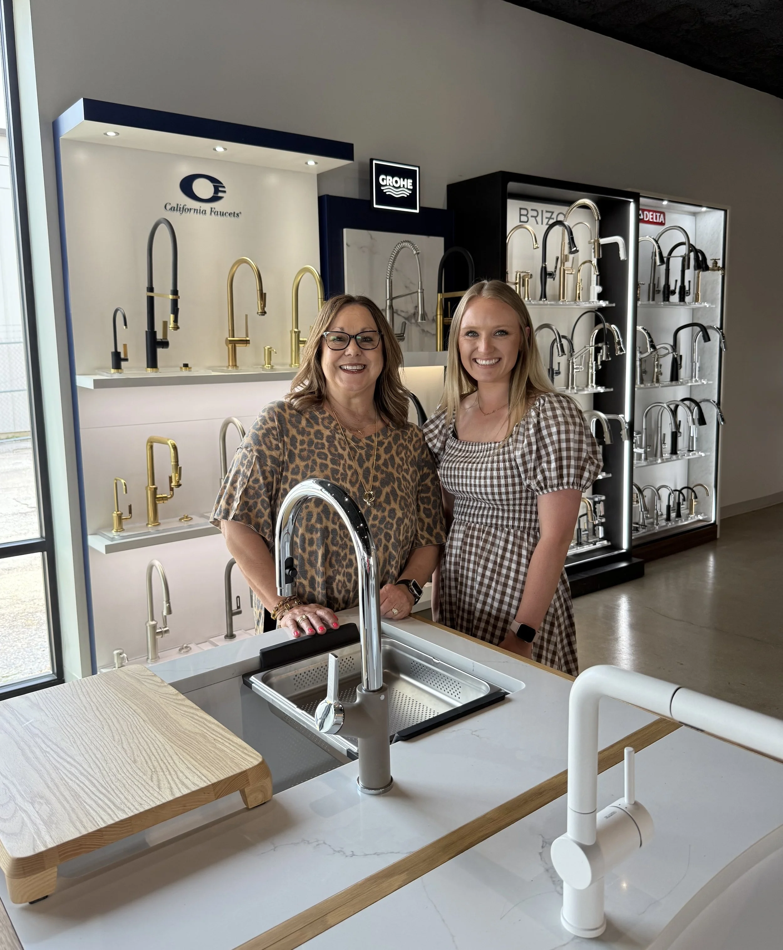 Two women are standing in front of a display of kitchen faucets on shelves in a showroom. The woman on the left has glasses and is wearing a leopard print top, and the woman on the right has blonde hair and is wearing a checkered dress. The display features various faucet styles in black, gold, and silver finishes.