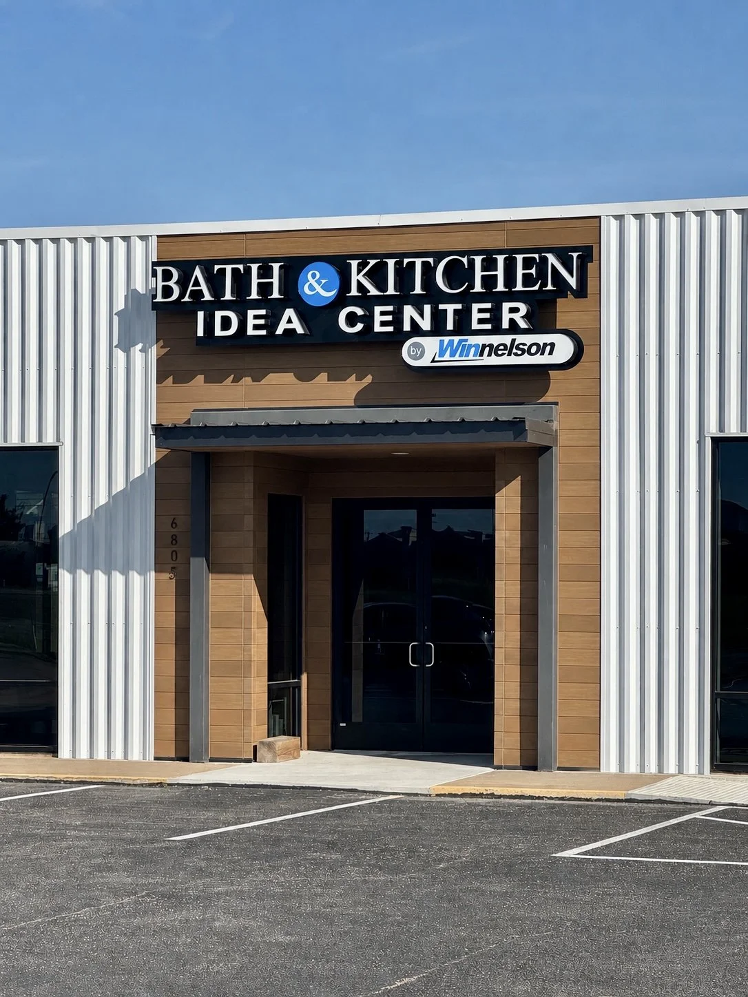 Exterior of a shop called Bath & Kitchen Idea Center by Winnelson, with a modern design featuring wood and metal siding, glass doors, and an empty parking lot in front.