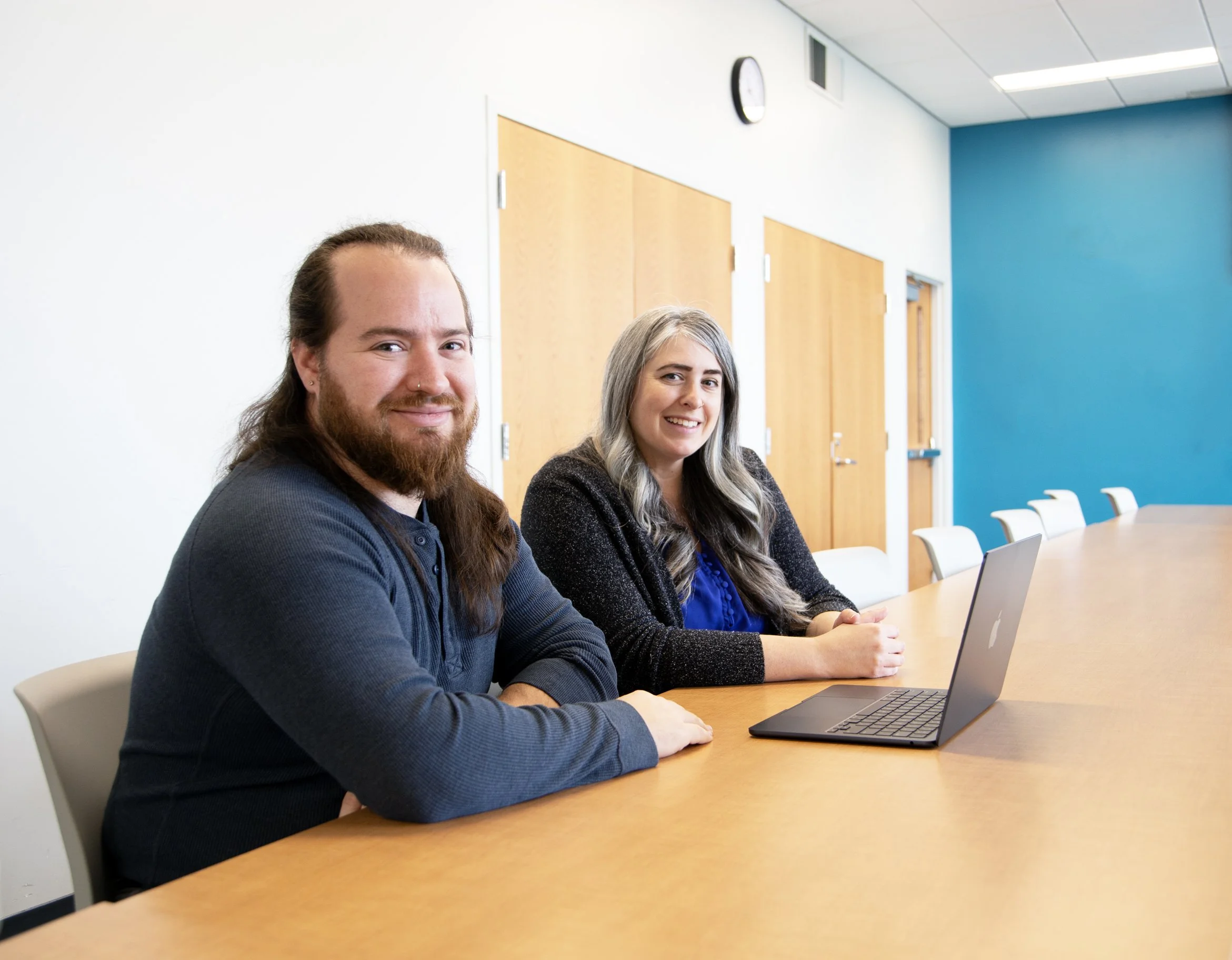 Two people sitting at a conference table with a laptop in front of them, smiling at the camera in a modern office room with a blue accent wall.