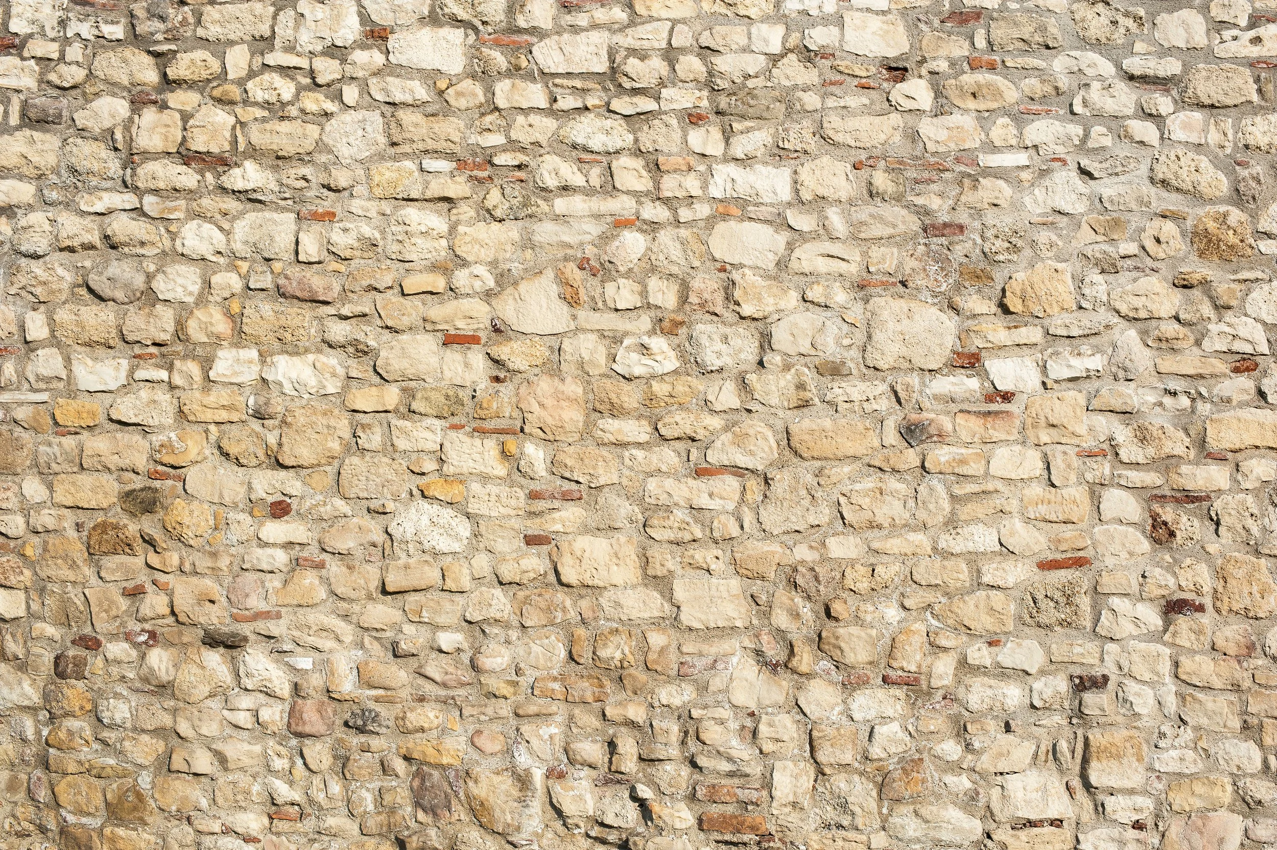 Mur en pierre beige avec différentes tailles de pierres et quelques petits accents rouges.