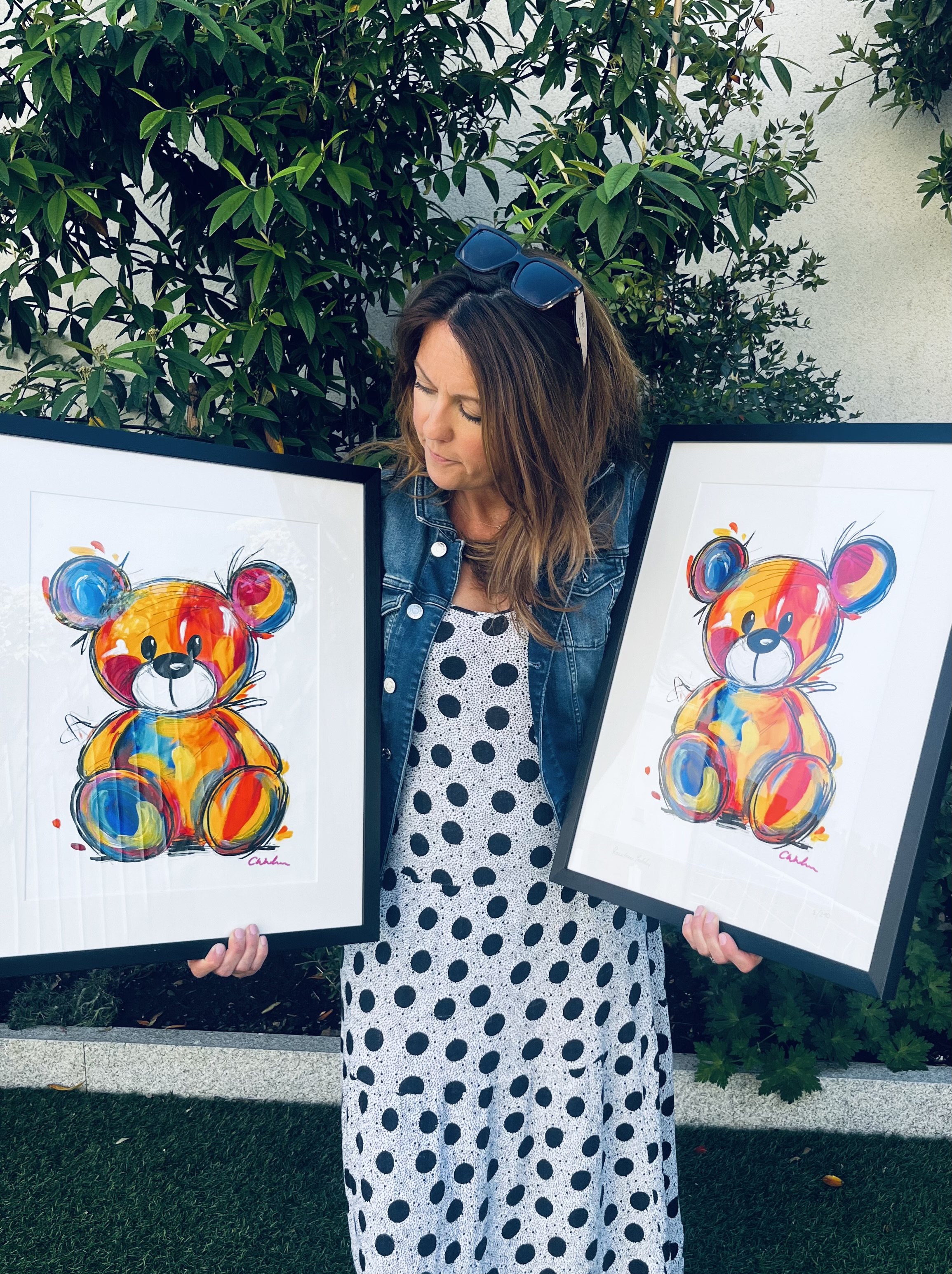 A woman standing outdoors in front of greenery, holding two framed colorful watercolor paintings of a bear. She has sunglasses on her head, is wearing a denim jacket, and a polka dot dress.