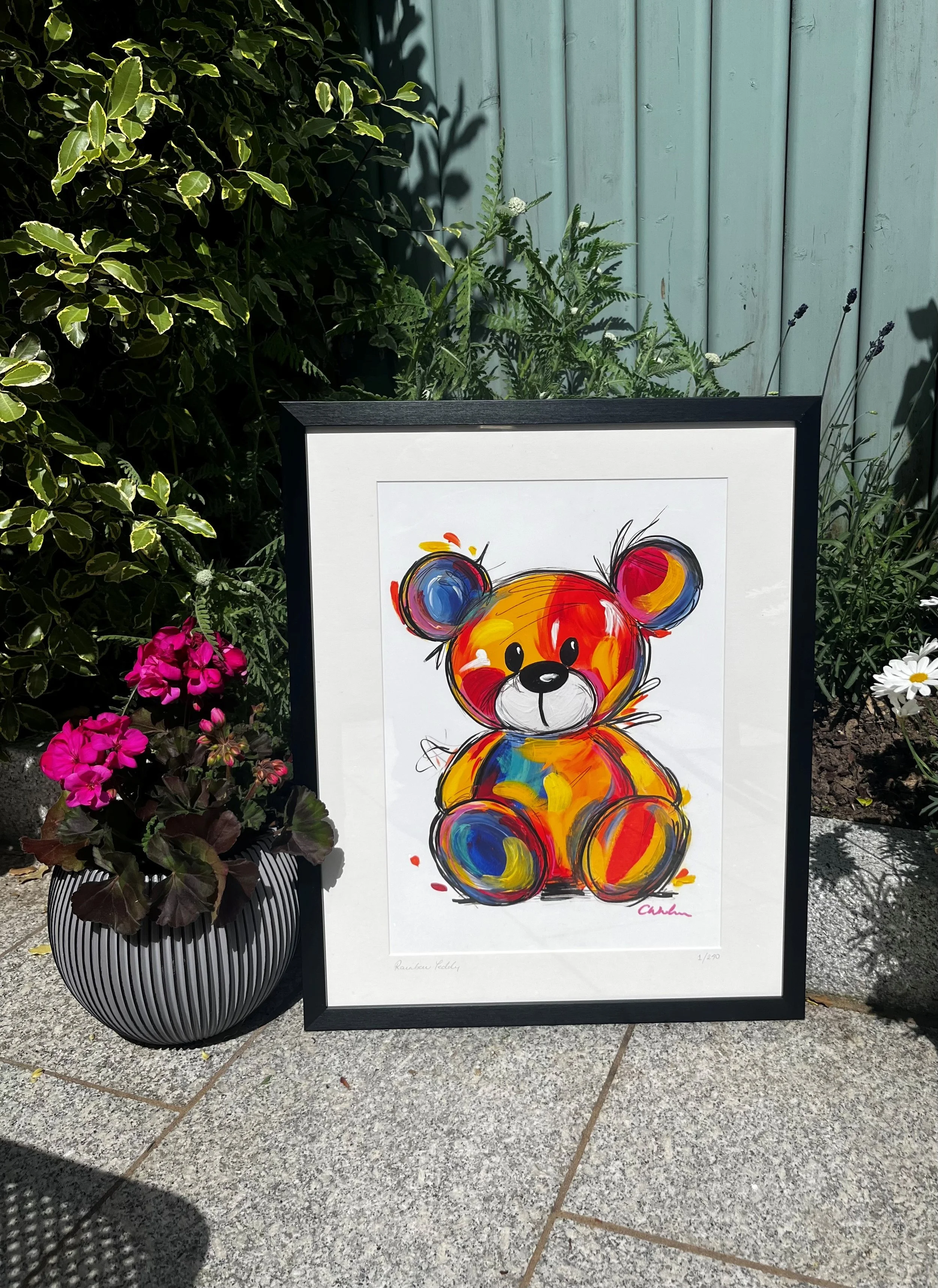 Colorful watercolor painting of a teddy bear with vibrant shades of red, yellow, blue, and black, framed and displayed outdoors with green foliage and flowers nearby.