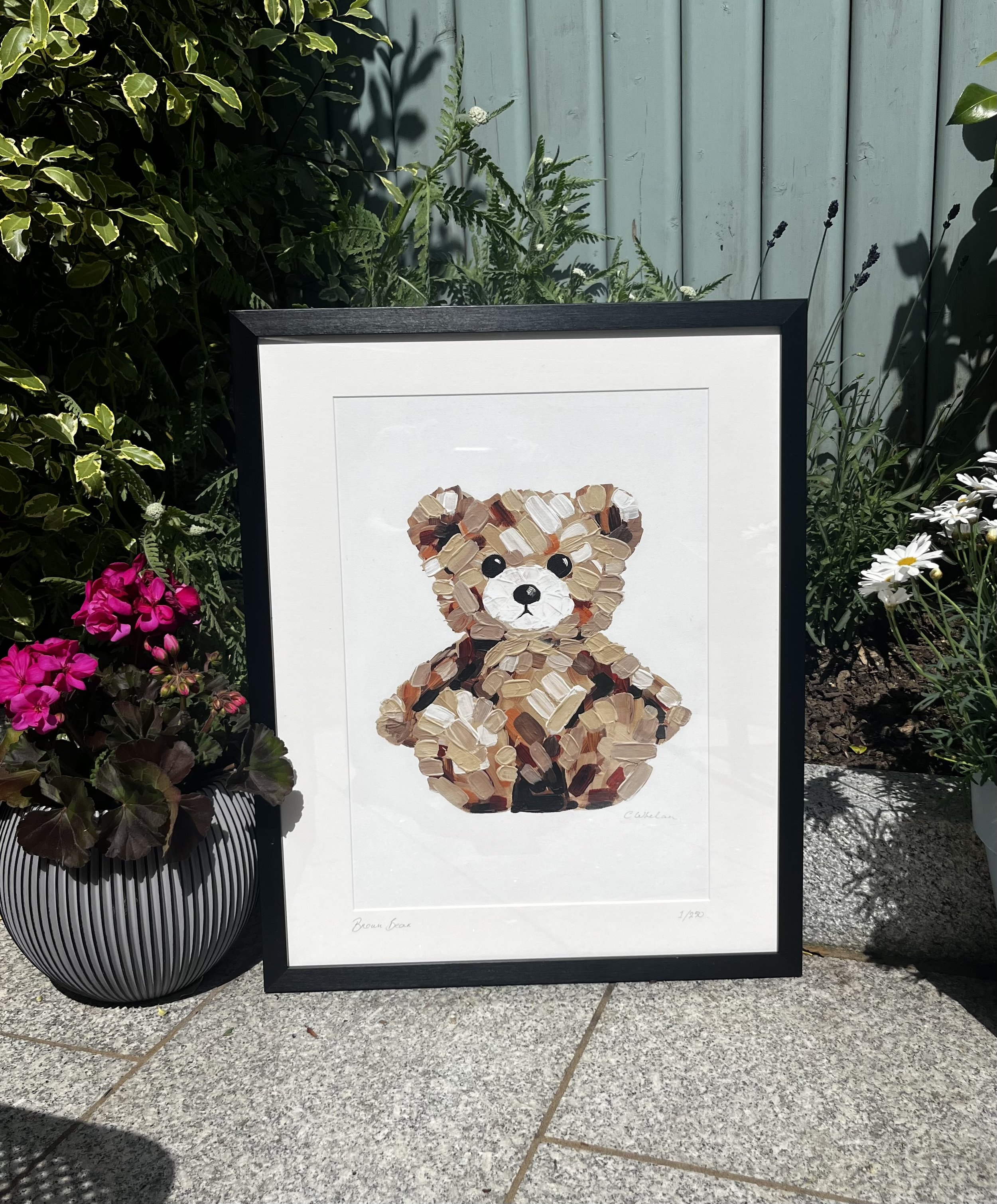 Framed artwork of a brown bear made with paintbrush strokes, placed outdoors on a tiled surface, with pink and purple flowers and greenery around.